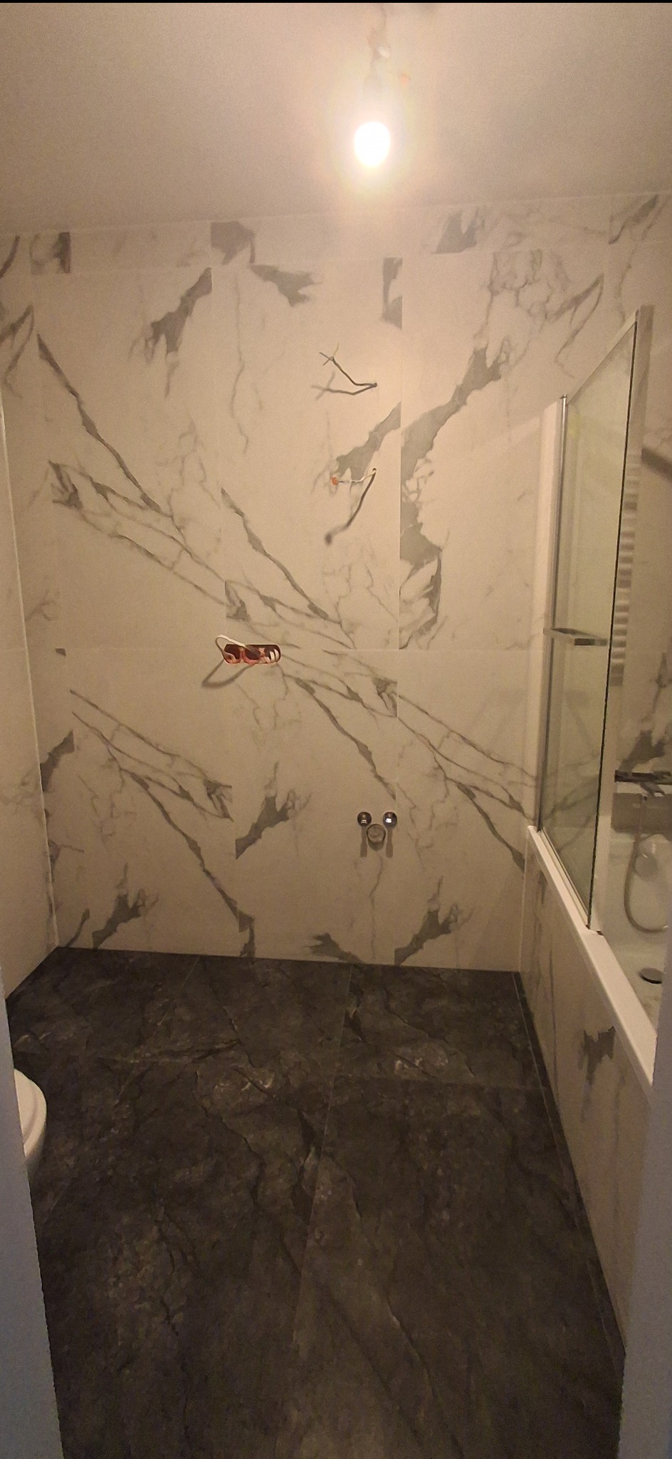 Wykończona łazienka z marmurowymi płytkami na ścianach i podłodze. Kabina prysznicowa ze szklanymi drzwiami. Widoczne wyprowadzone kable elektryczne.