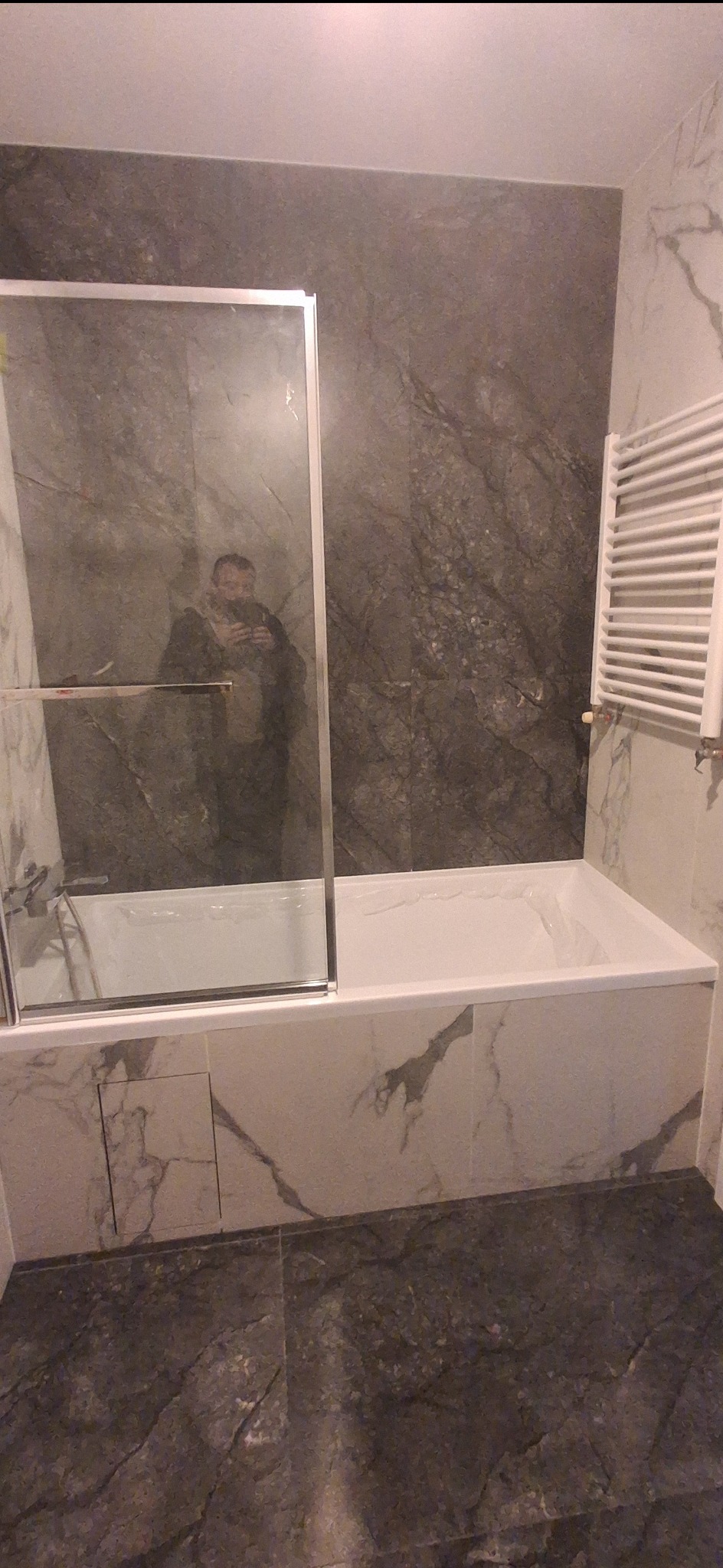 Wykończona łazienka z wanną i szklanym parawanem, płytki imitujące marmur na ścianach i podłodze, grzejnik drabinkowy. Widoczne odbicie osoby robiącej zdjęcie.