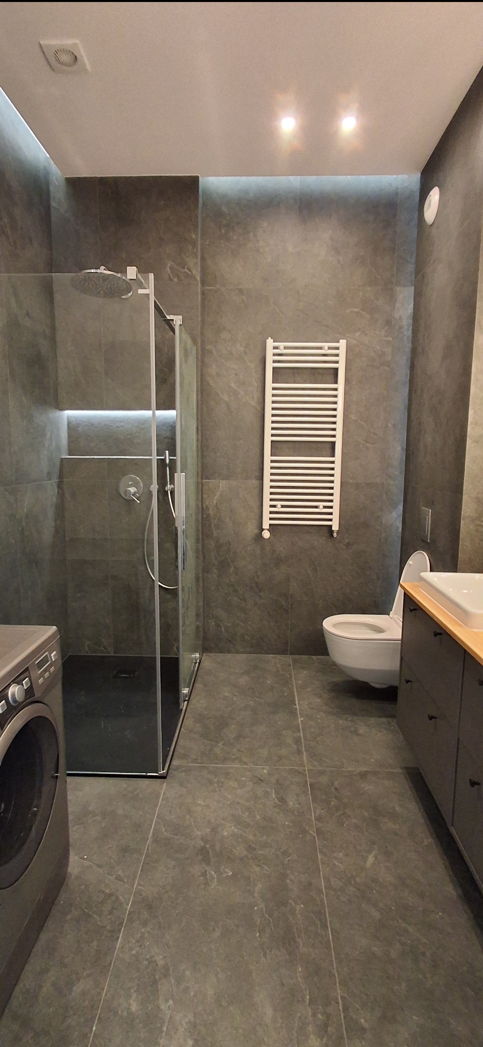 Nowoczesna łazienka z kabiną prysznicową typu walk-in, grzejnikiem drabinkowym i umywalką z szafką w odcieniach szarości. Minimalistyczny design z akcentami światła.