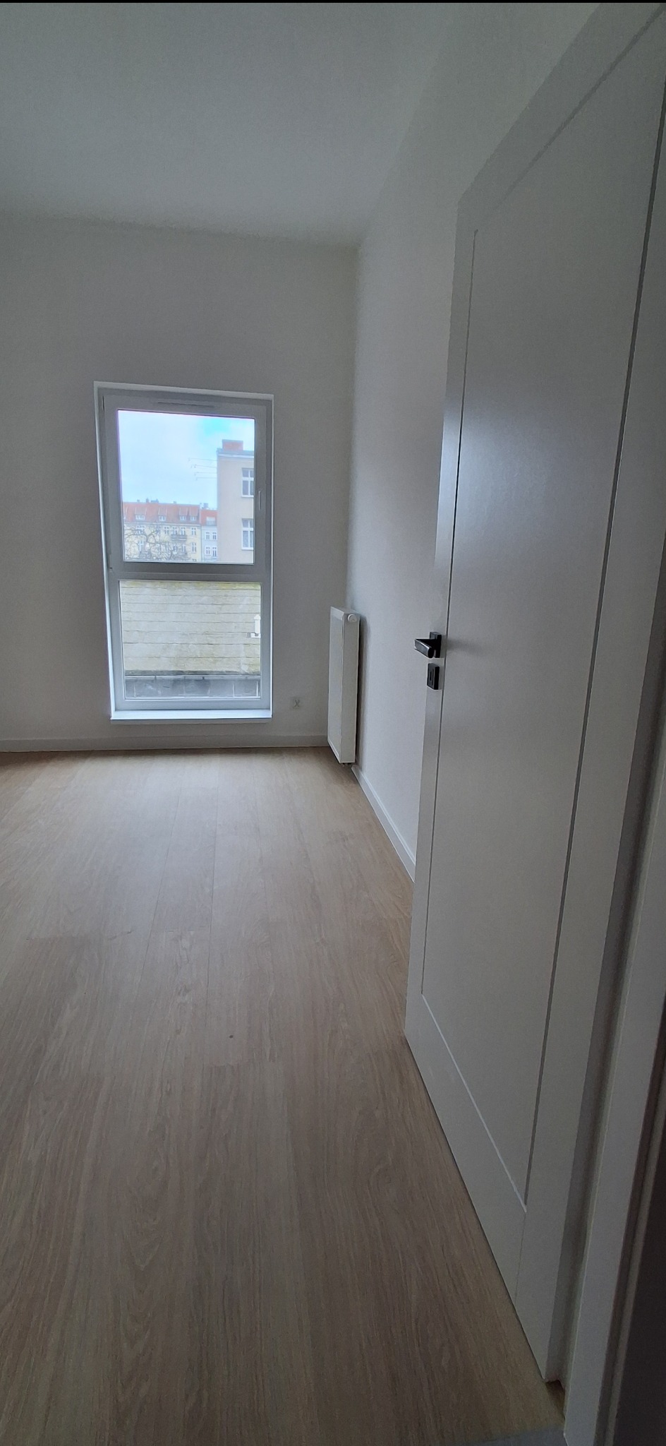 Jasne panele podłogowe w pustym pokoju z białymi ścianami, oknem i drzwiami. Widoczny grzejnik. Minimalistyczne wykończenie wnętrza.