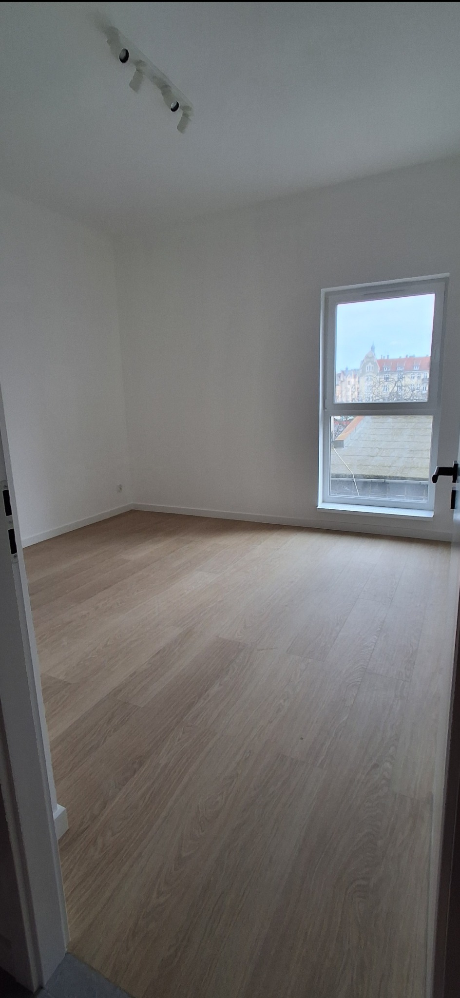 Jasne panele podłogowe w pustym pokoju z białymi ścianami i oknem z widokiem na miasto. Minimalistyczne oświetlenie sufitowe.