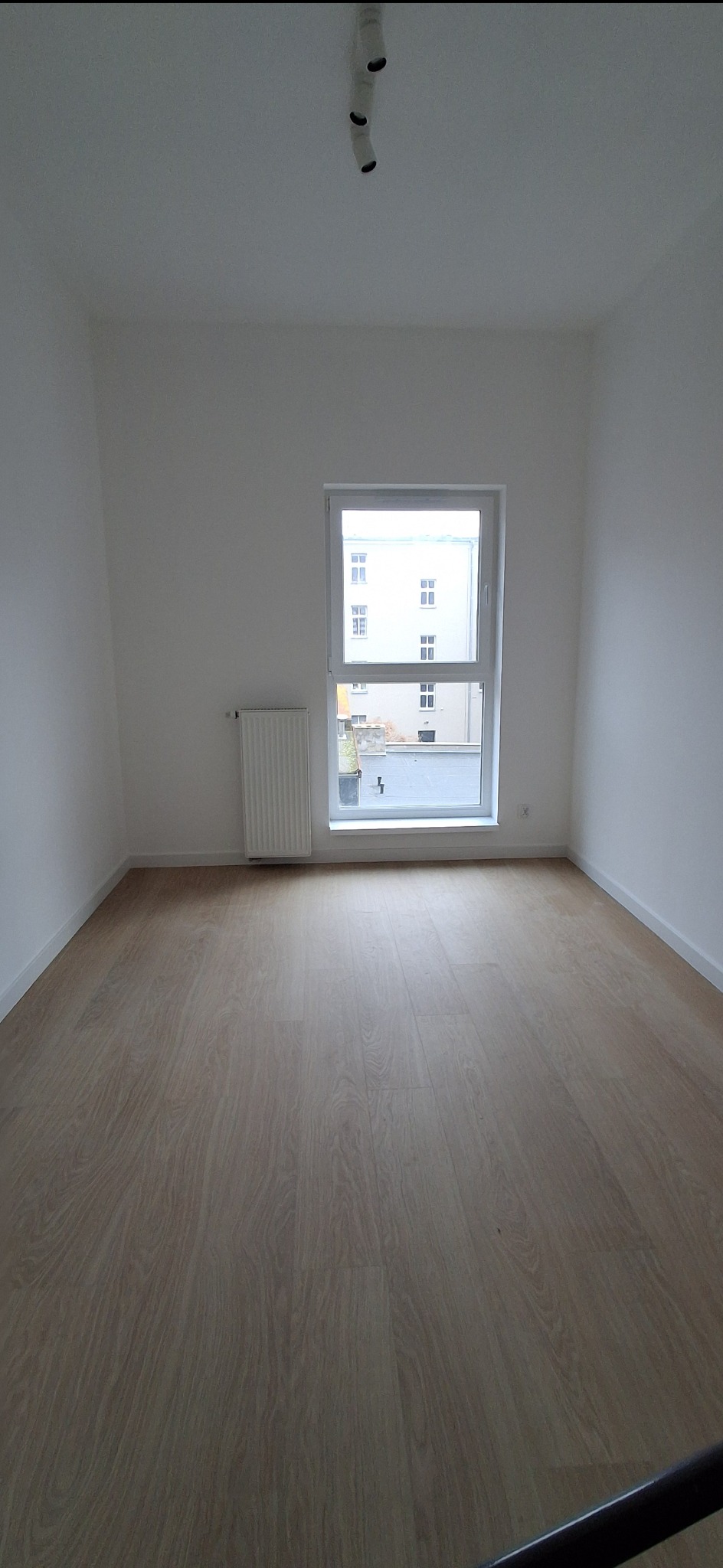 Jasne panele podłogowe w pustym, pomalowanym na biało pokoju z oknem i grzejnikiem. Minimalistyczne wnętrze z trzema punktami świetlnymi na suficie.