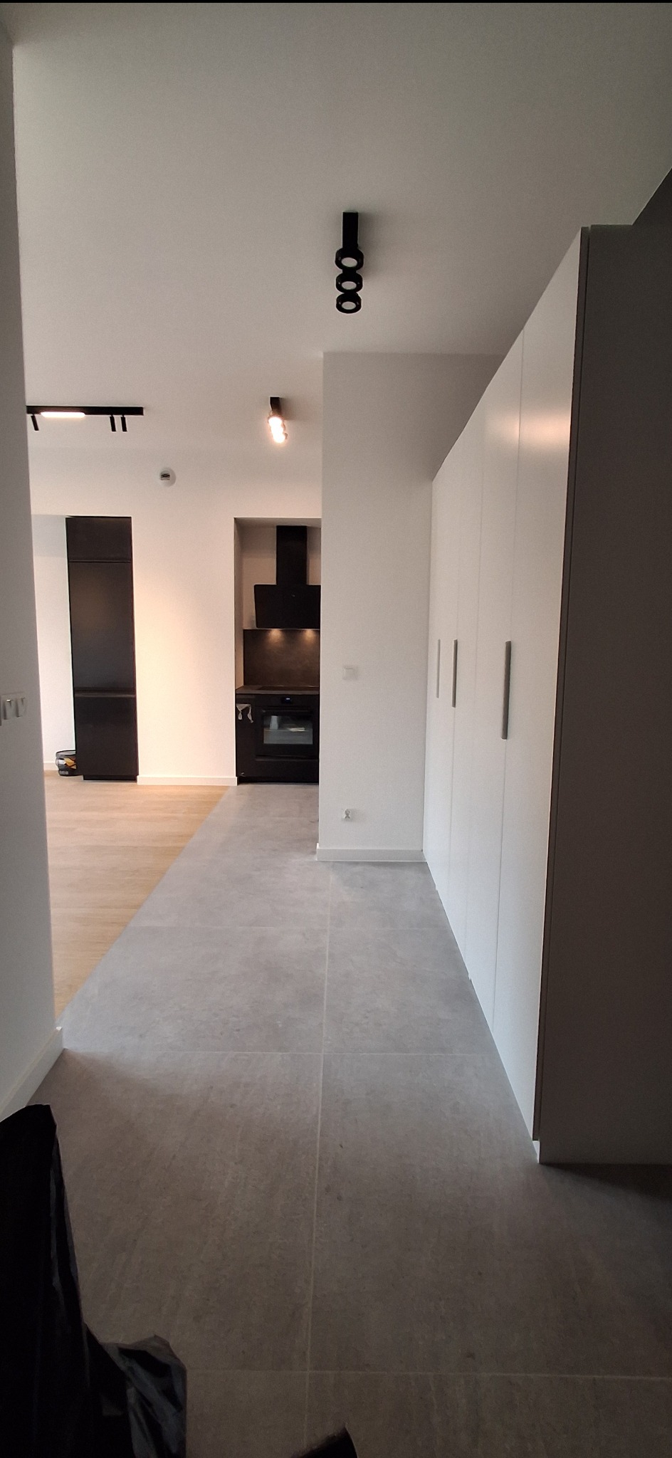 Nowoczesne wnętrze mieszkania z szarą podłogą, białą zabudową i czarnymi akcentami oświetleniowymi. Widok na kuchnię z czarnym okapem i sprzętem.