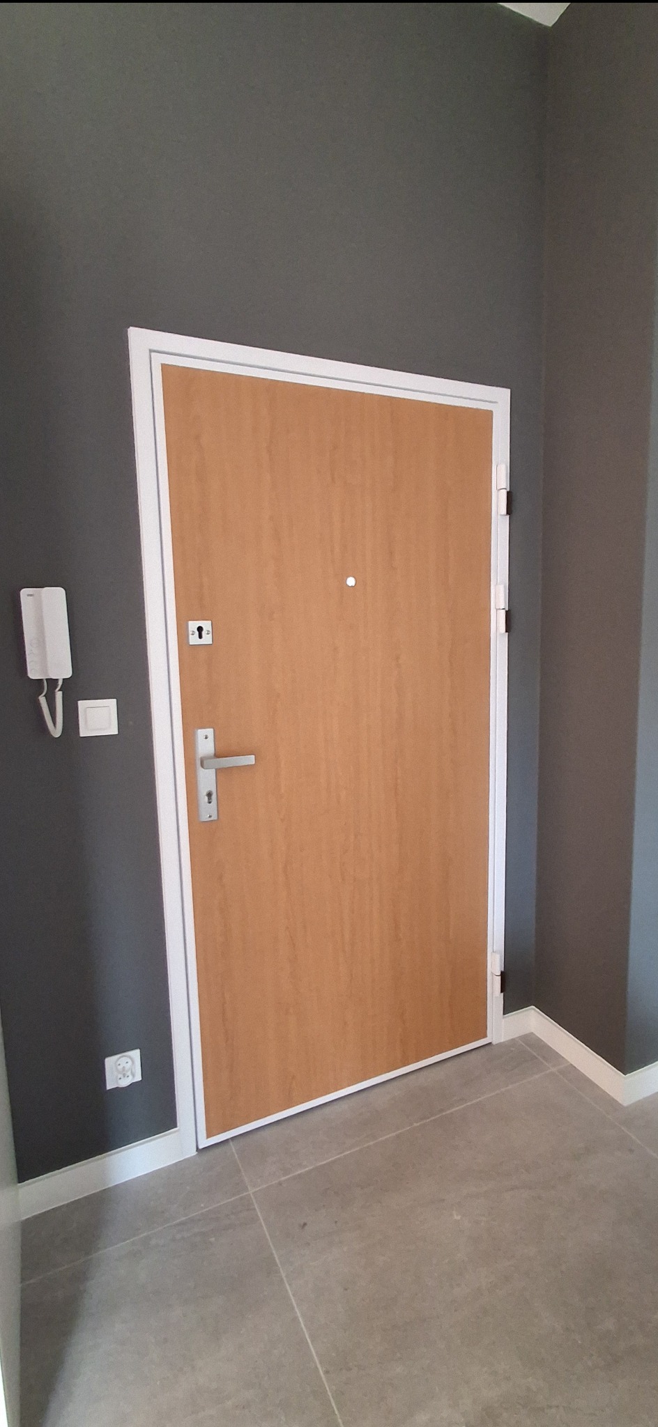 Zamknięte, drewniane drzwi wejściowe z wizjerem i srebrną klamką, osadzone w białej futrynie. Szare ściany i podłoga z płytek. Widoczny domofon.