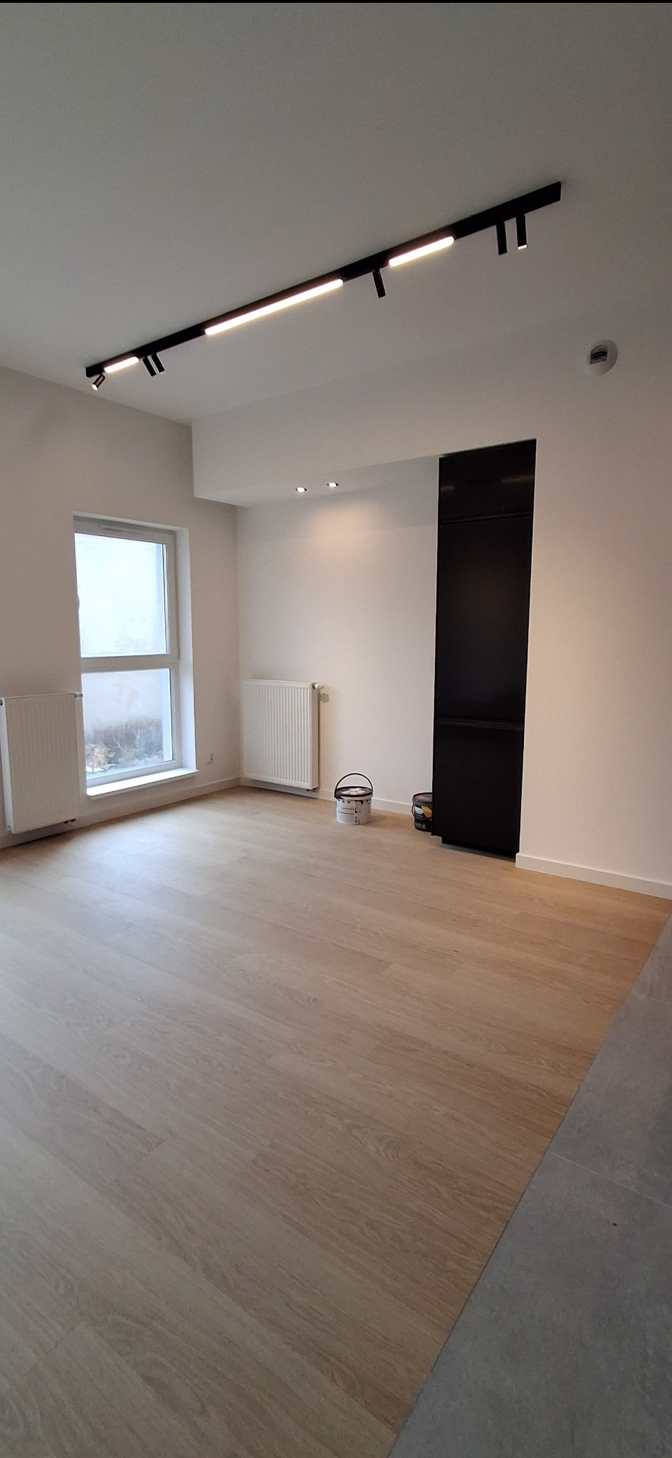 Jasne panele podłogowe w nowym wnętrzu z grzejnikami i nowoczesnym oświetleniem sufitowym. Widoczne wiaderka po farbie.