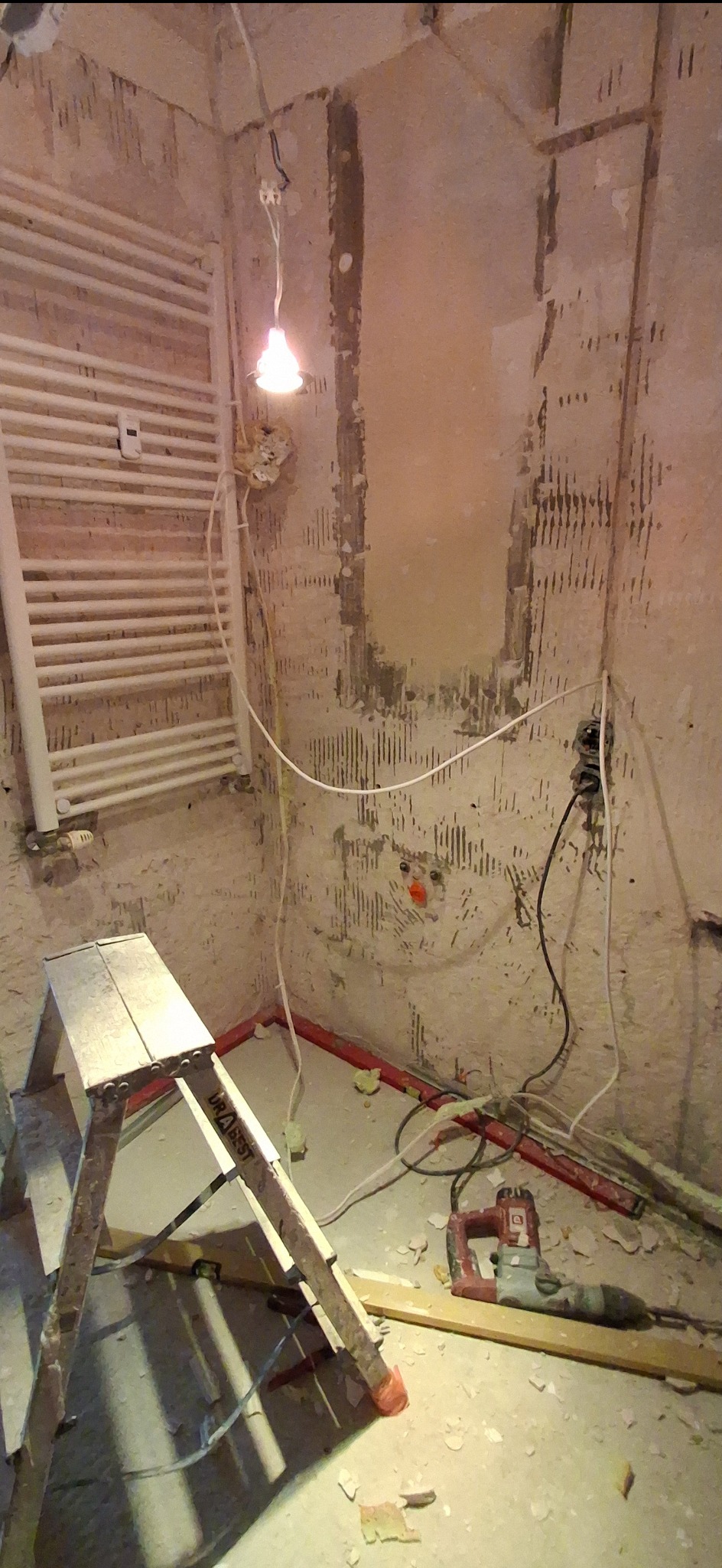 Wnętrze łazienki w trakcie demontażu: widoczne ślady po płytkach, grzejnik drabinkowy, przewody elektryczne, drabina i młot udarowy na podłodze.