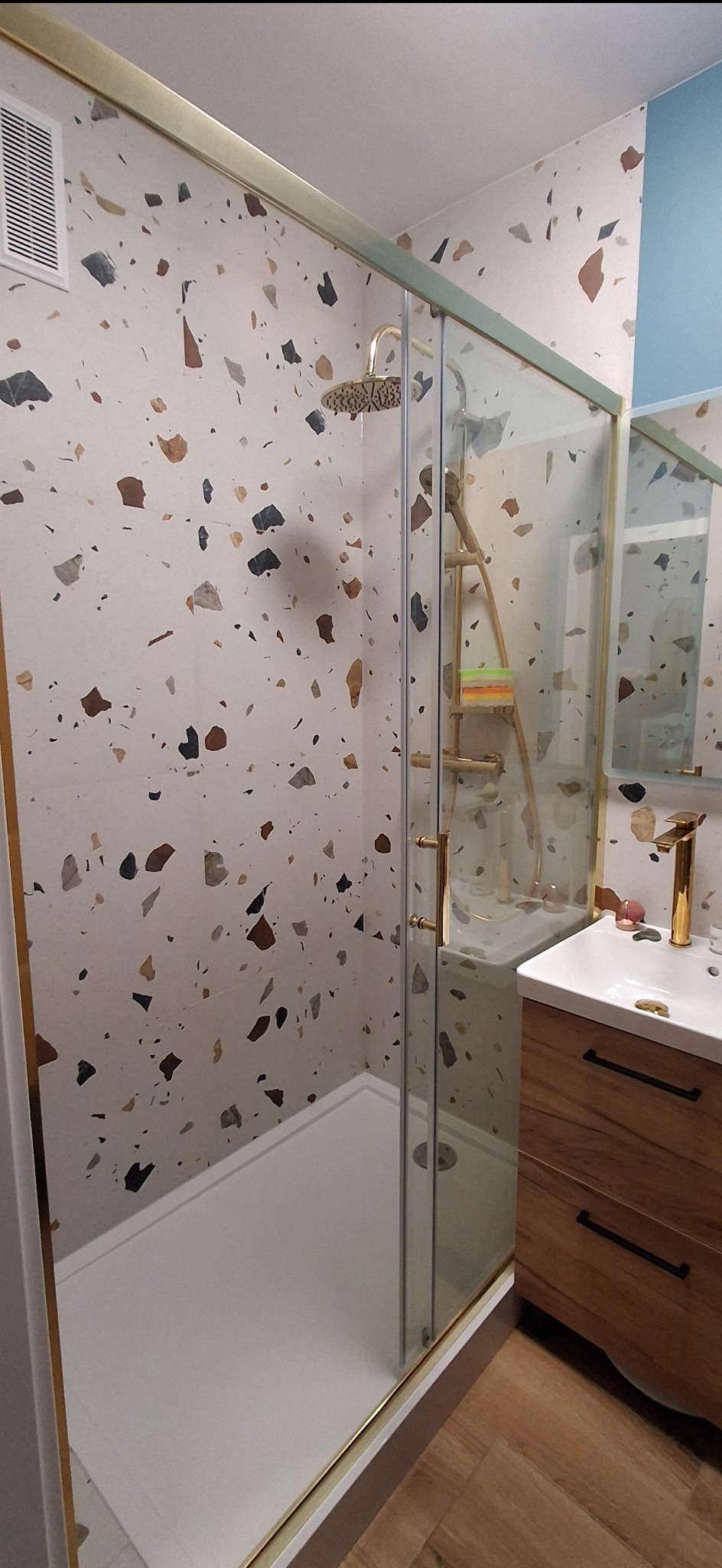 Nowoczesna kabina prysznicowa z deszczownicą i złotymi detalami, umywalka z szafką z drewna. Ściany wykończone płytkami w stylu lastryko.