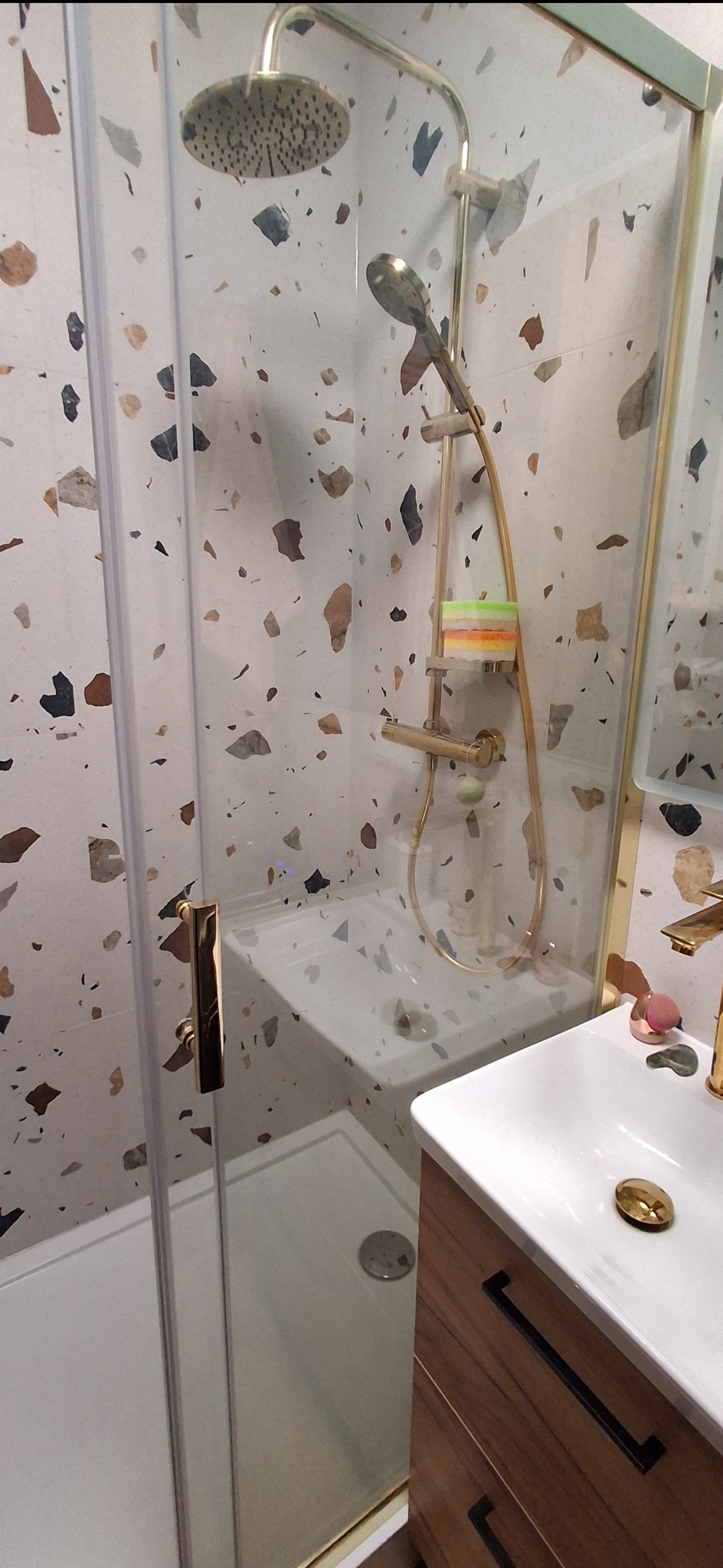 Nowoczesna kabina prysznicowa z deszczownicą i złotą armaturą na tle ściany z mozaiką w odcieniach brązu i beżu. Widoczna umywalka z szafką.