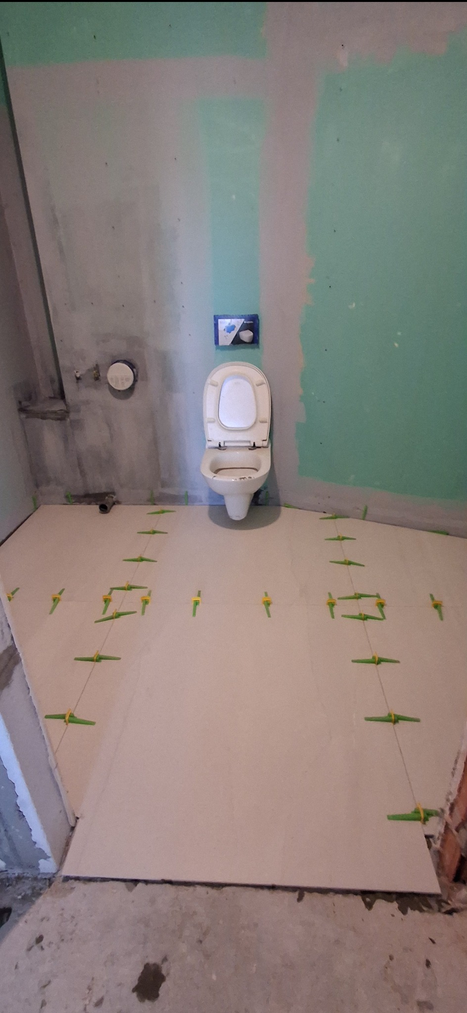 Białe płytki podłogowe w trakcie układania w łazience, z zielonymi krzyżykami dystansowymi. Ściany częściowo pokryte płytą gipsową, widoczna toaleta i instalacja.