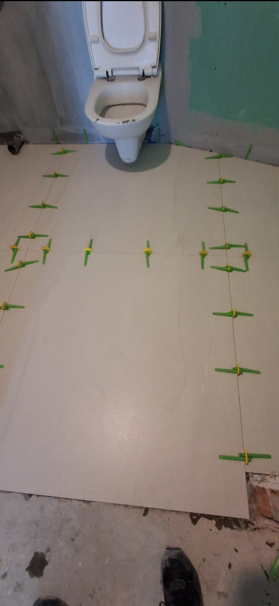 Układanie jasnych płytek podłogowych w łazience, widoczne krzyżyki dystansowe i częściowo zamontowana toaleta. Etap prac wykończeniowych.