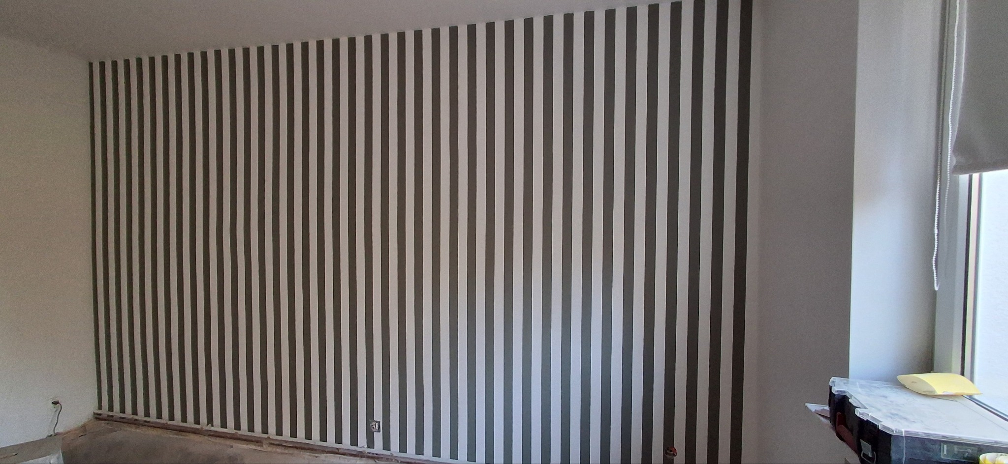 Ściana w pokoju po malowaniu w pionowe, szare pasy. Widoczne narzędzia malarskie przy oknie. Wykończenie wnętrz w nowoczesnym stylu.