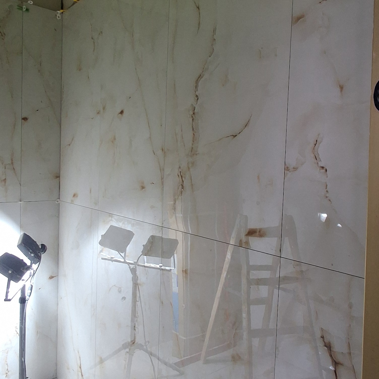 Wykończona ściana w łazience z dużymi, marmurkowymi płytkami. Widoczne oświetlenie budowlane i drabina. Płytki ułożone z precyzją, z zachowaniem równych fug.