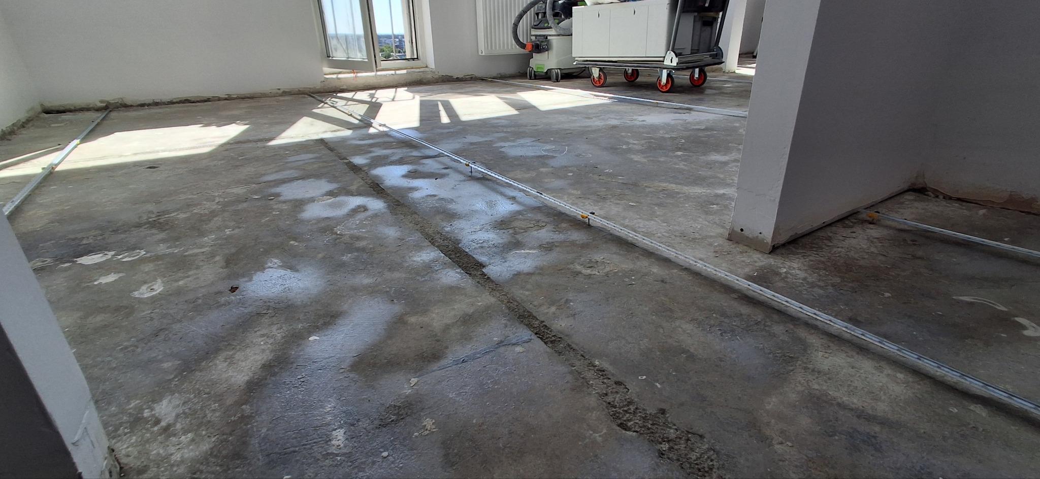 Przygotowanie podłoża pod wylewkę samopoziomującą. Widoczne listwy poziomujące i ślady po wcześniejszych pracach. W tle okno i sprzęt budowlany.