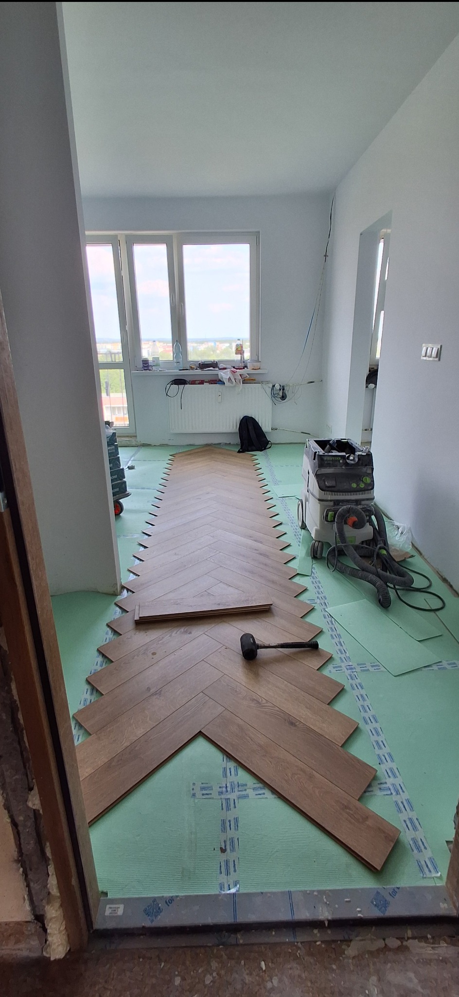 Układanie paneli w jodełkę na podkładzie. Widoczny gumowy młotek i odkurzacz przemysłowy. Jasne wnętrze z oknem i widokiem na miasto.