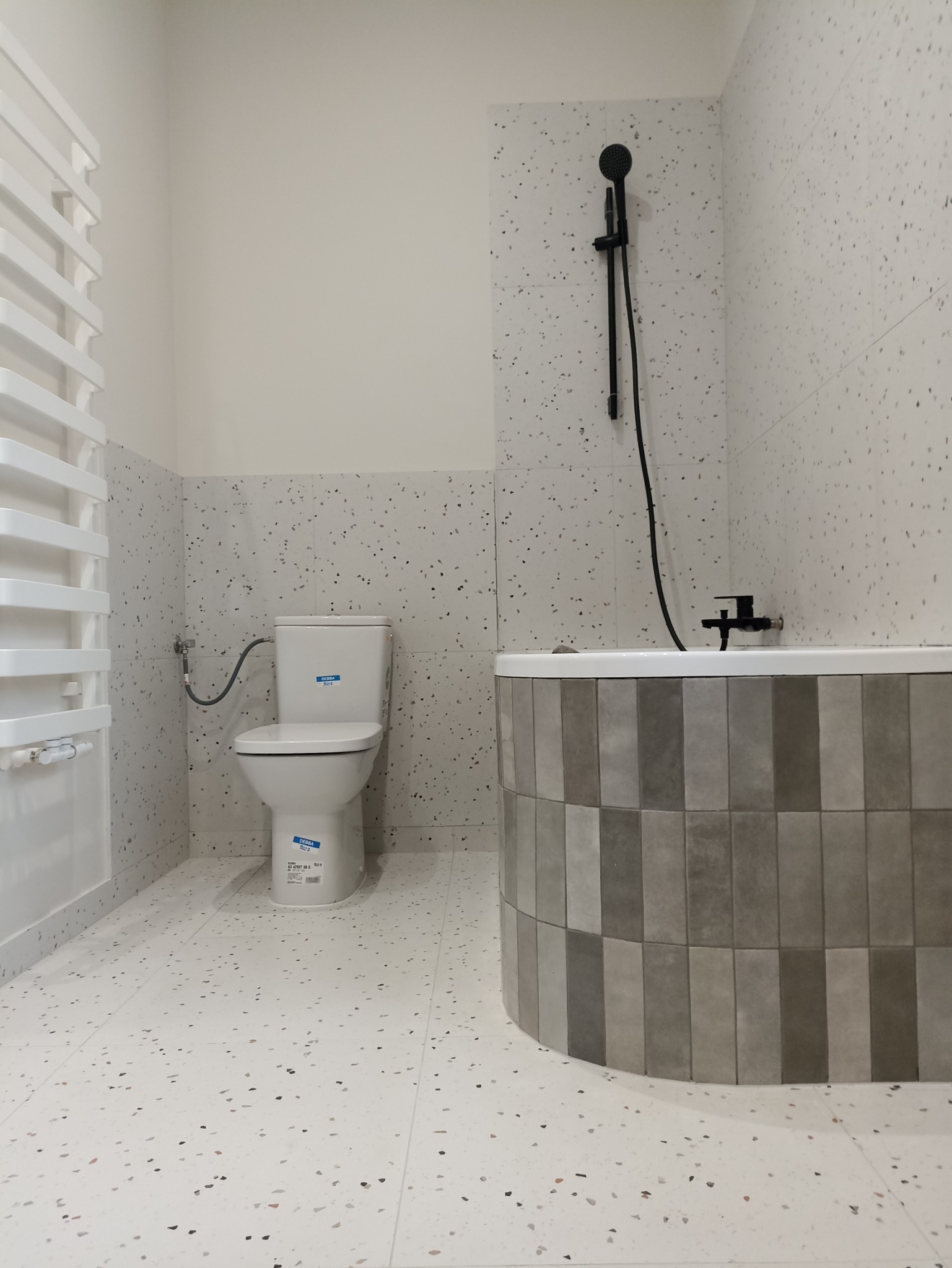 Nowoczesna łazienka z toaletą, grzejnikiem drabinkowym i obudowaną wanną w szaro-białej kolorystyce z terazzo na podłodze i ścianach.