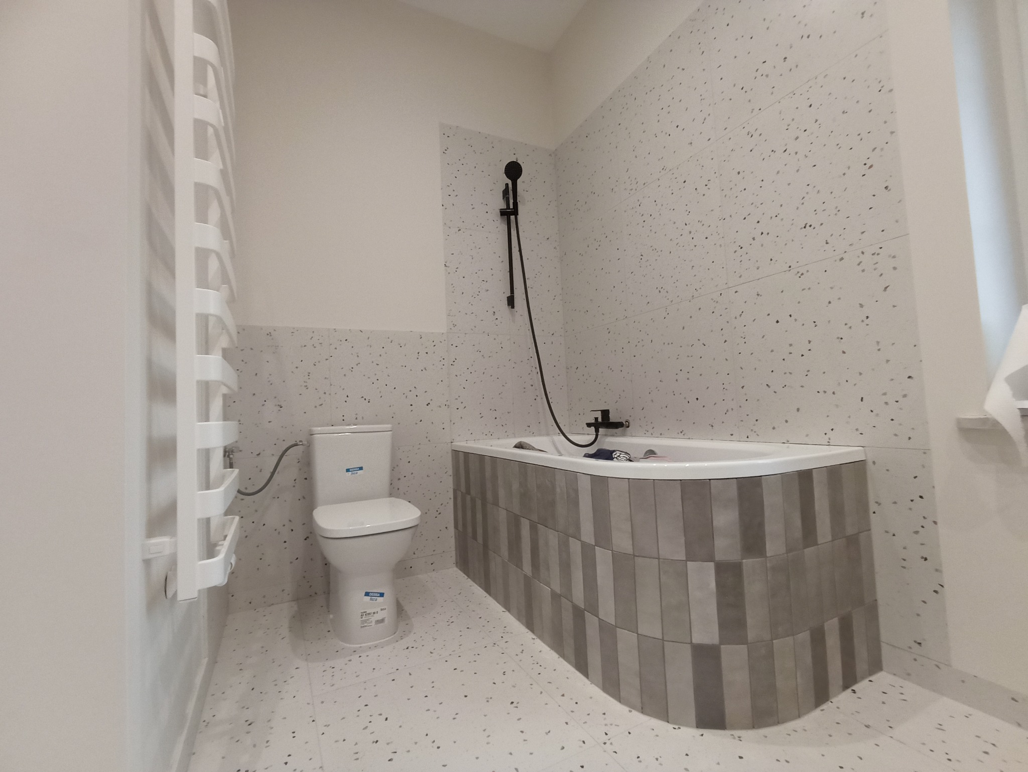 Nowoczesna łazienka po remoncie w Suchym Lesie, z wanną obudowaną szarymi płytkami w pionowy wzór, toaletą i grzejnikiem drabinkowym.