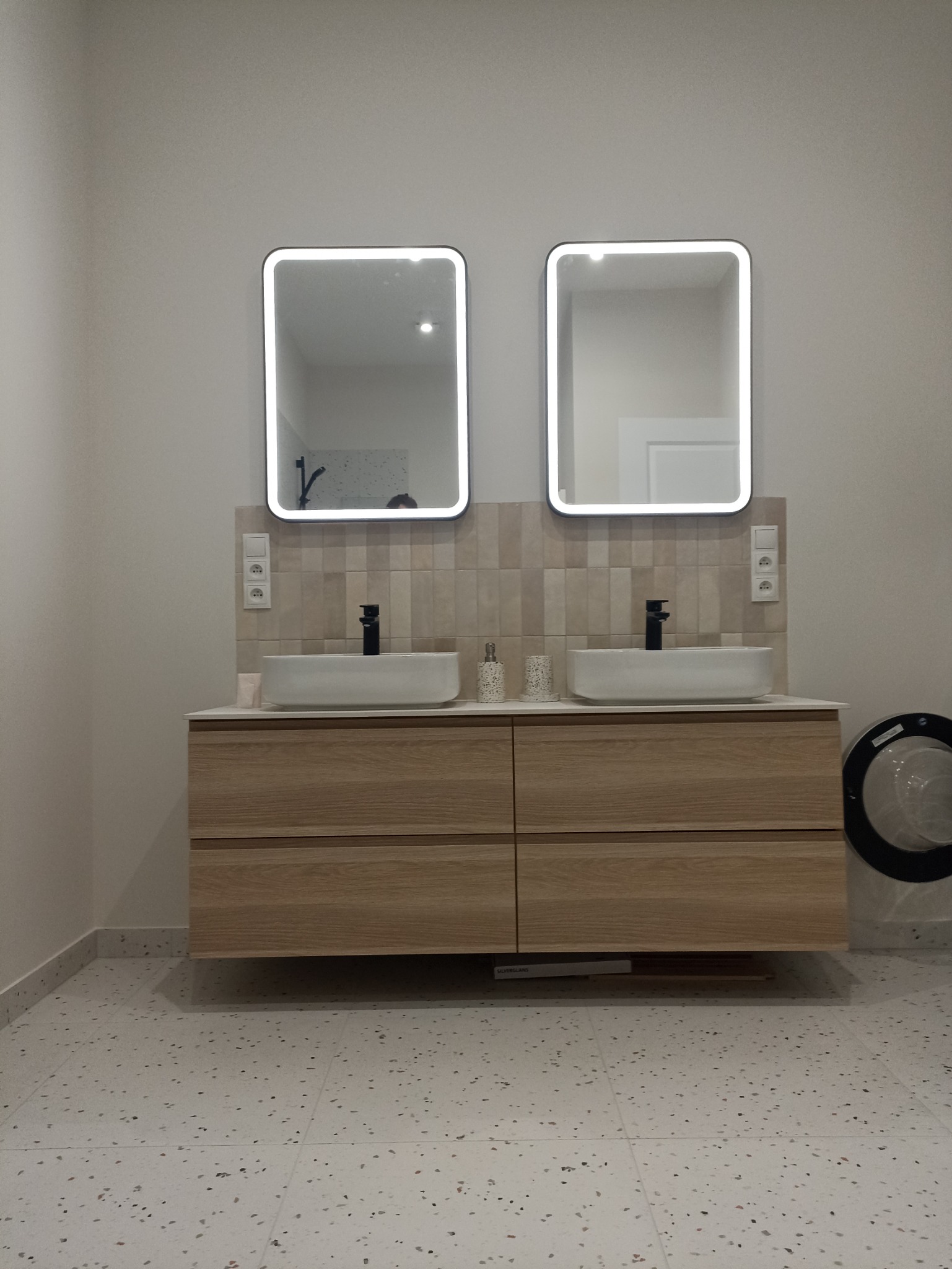 Nowoczesna łazienka z dwoma umywalkami nablatowymi, podświetlanymi lustrami i drewnianą szafką z szufladami, na podłodze płytki lastryko, widoczna pralka z czarnym obramowaniem.