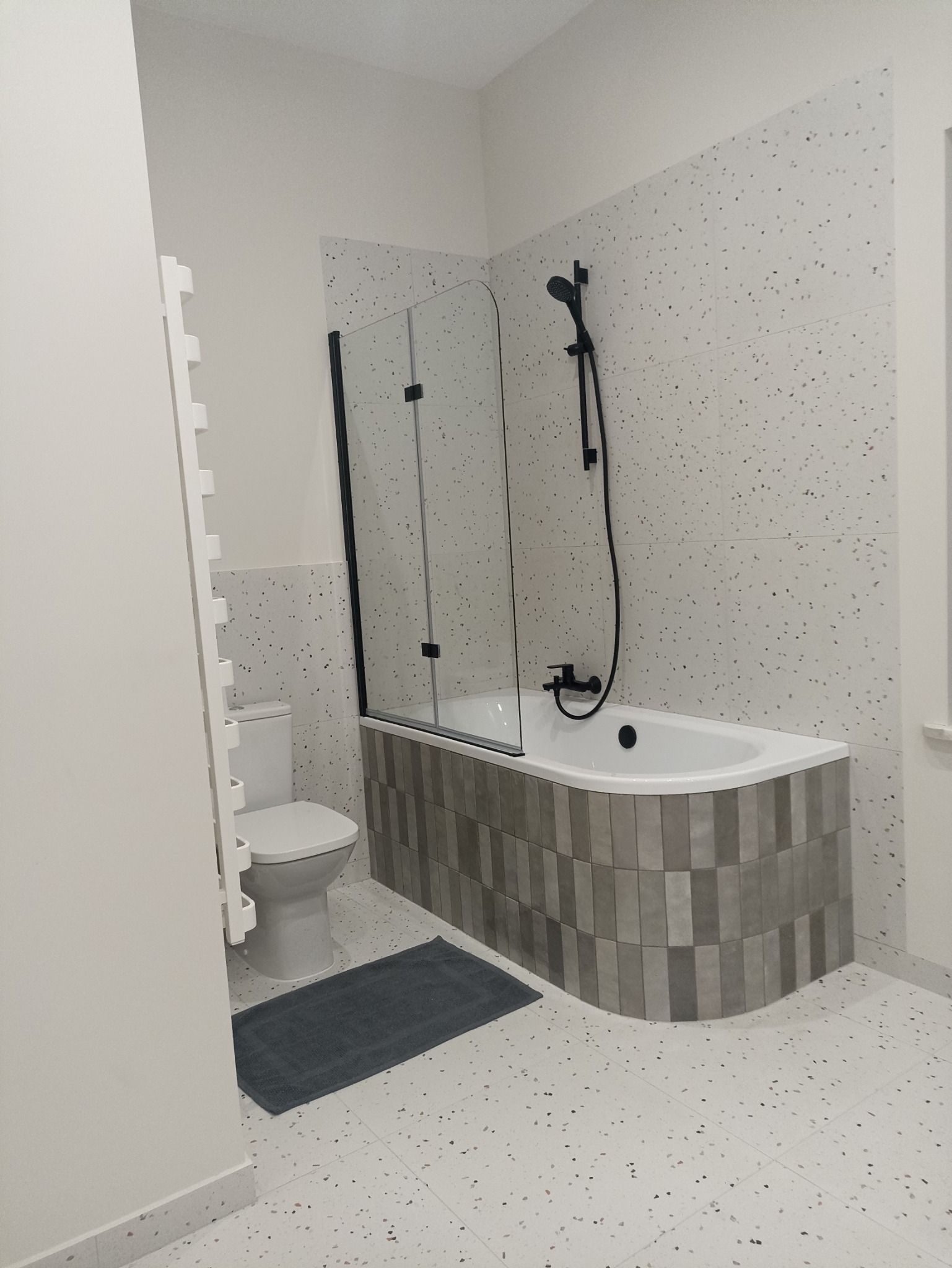 Nowoczesna łazienka po renowacji z wanną obudowaną szarymi płytkami, czarną armaturą i szklanym parawanem, podłoga i ściany wyłożone płytkami imitującymi lastryko.