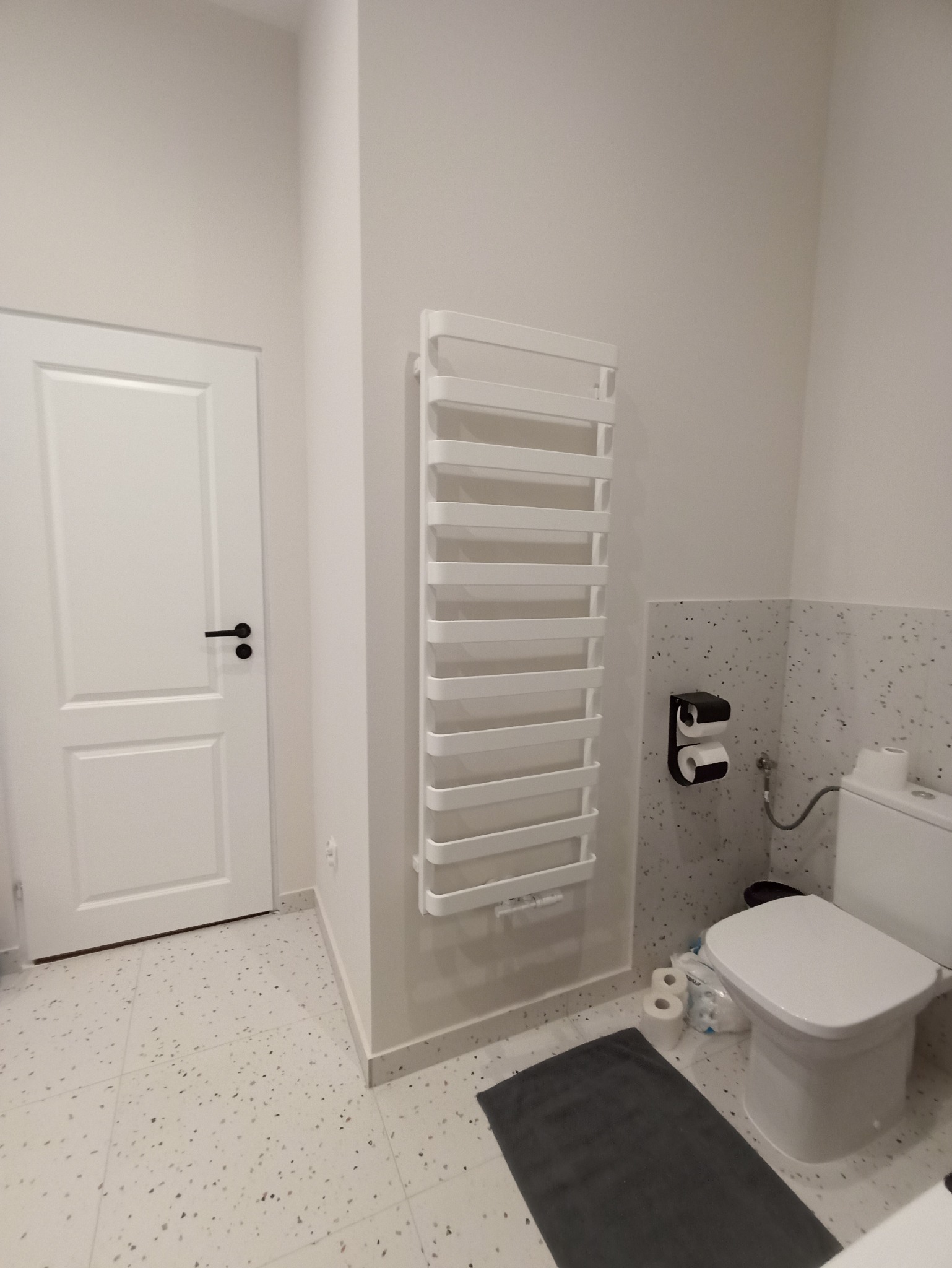 Jasna łazienka po remoncie z białymi płytkami lastryko na podłodze i ścianie, grzejnikiem drabinkowym, białą toaletą i drzwiami z czarną klamką.