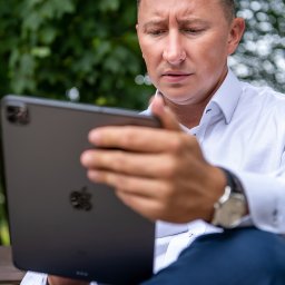 FINDOM Kancelaria Kredytów Hipotecznych Jarosław Fuzowski - Mężczyzna w białej koszuli i granatowych spodniach, siedząc na ławce w parku, z zamyśloną miną przegląda coś na tablecie.