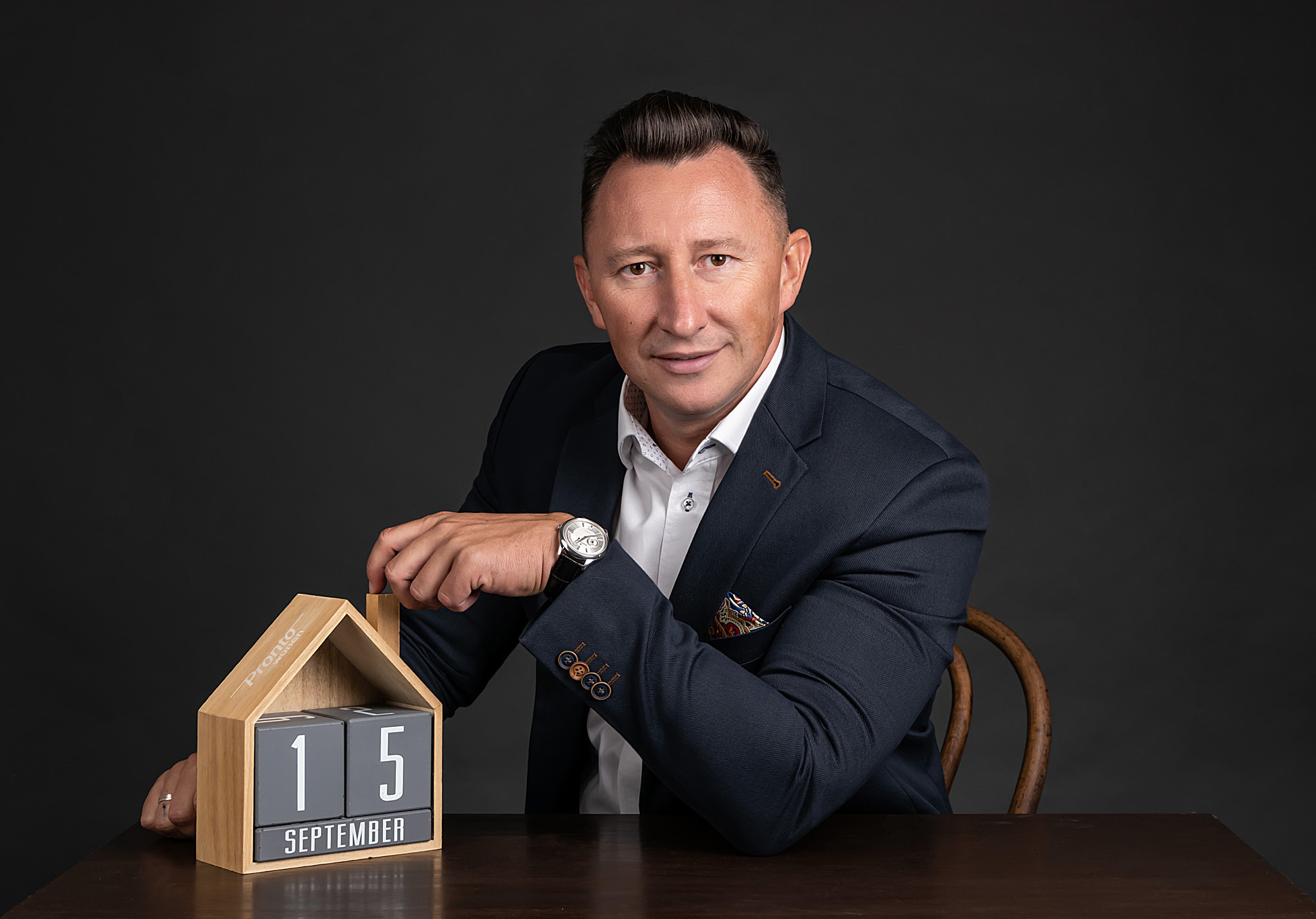 Mężczyzna w garniturze siedzi za biurkiem, obok niego drewniany kalendarz w kształcie domu wskazujący datę 15 września.