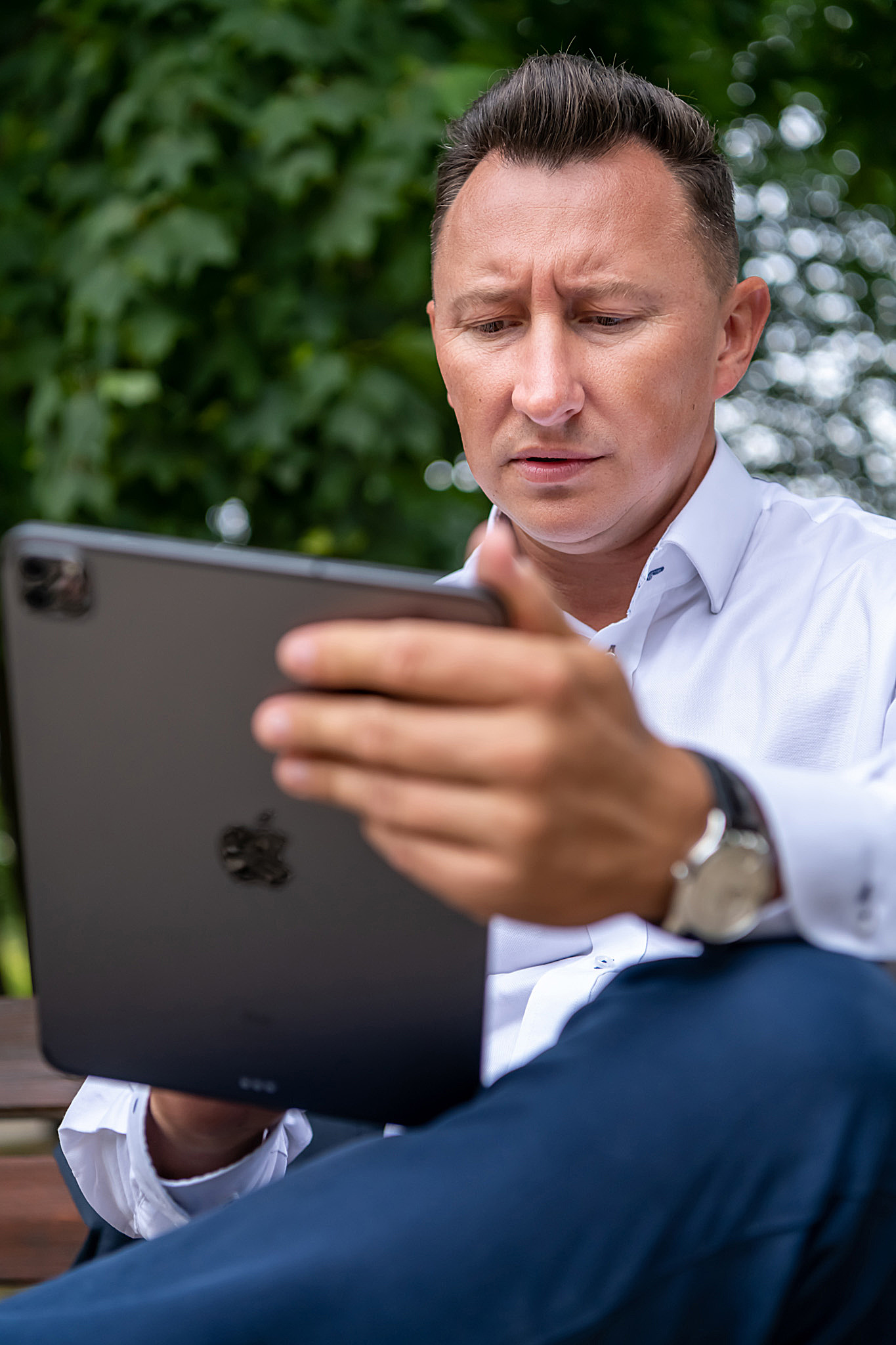 Mężczyzna w białej koszuli i granatowych spodniach, siedząc na ławce w parku, z zamyśloną miną przegląda coś na tablecie.