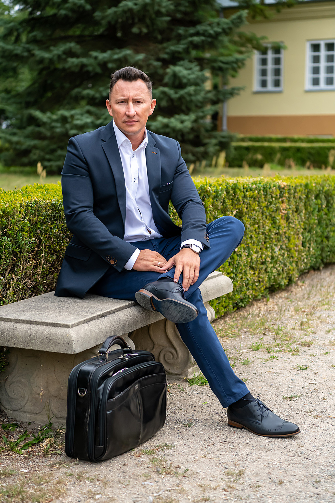 Mężczyzna w garniturze siedzi na kamiennej ławce w parku, obok leży czarna skórzana aktówka, w tle żywopłot i budynek z białymi oknami.