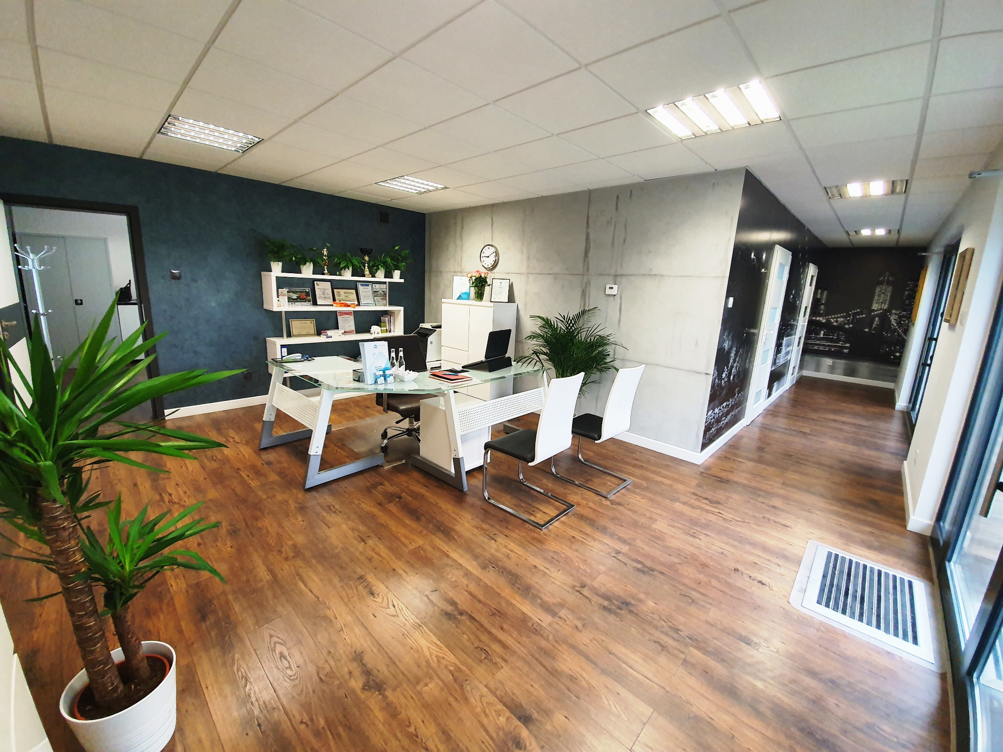 Jasne biuro z drewnianą podłogą, szklanym biurkiem, białymi krzesłami, roślinami doniczkowymi i dekoracyjną ścianą z motywem miasta.