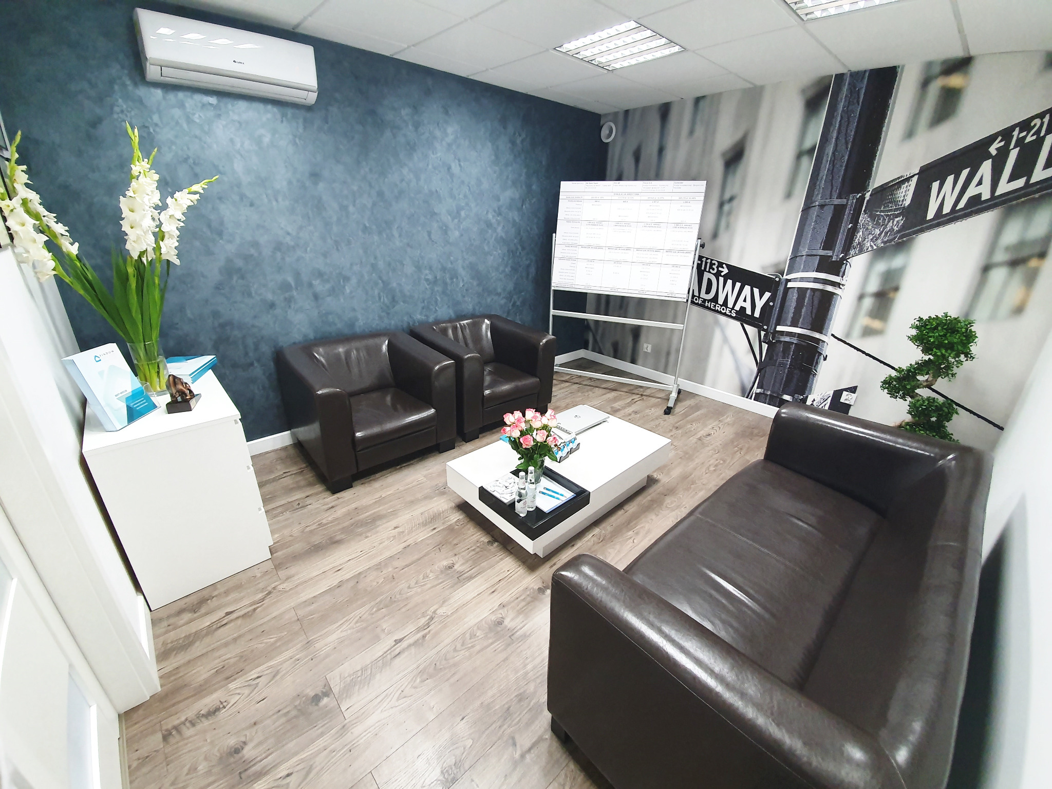 Eleganckie biuro z ciemnobrązowymi skórzanymi fotelami i kanapą, białą szafką i stolikiem kawowym z kwiatami, tablicą suchościeralną z danymi i tapetą z motywem ulicy Wall Street.