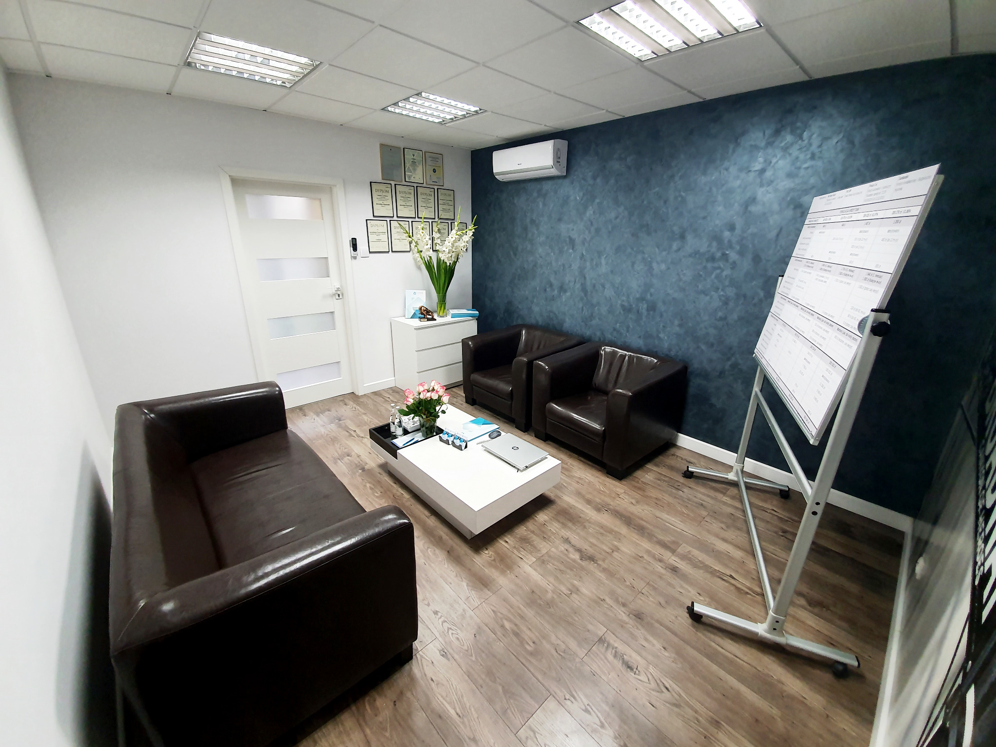 Wnętrze biura doradcy kredytowego: brązowe sofy, biały stolik kawowy z laptopem i kwiatami, tablica suchościeralna z danymi, dyplomy na ścianie, klimatyzacja.