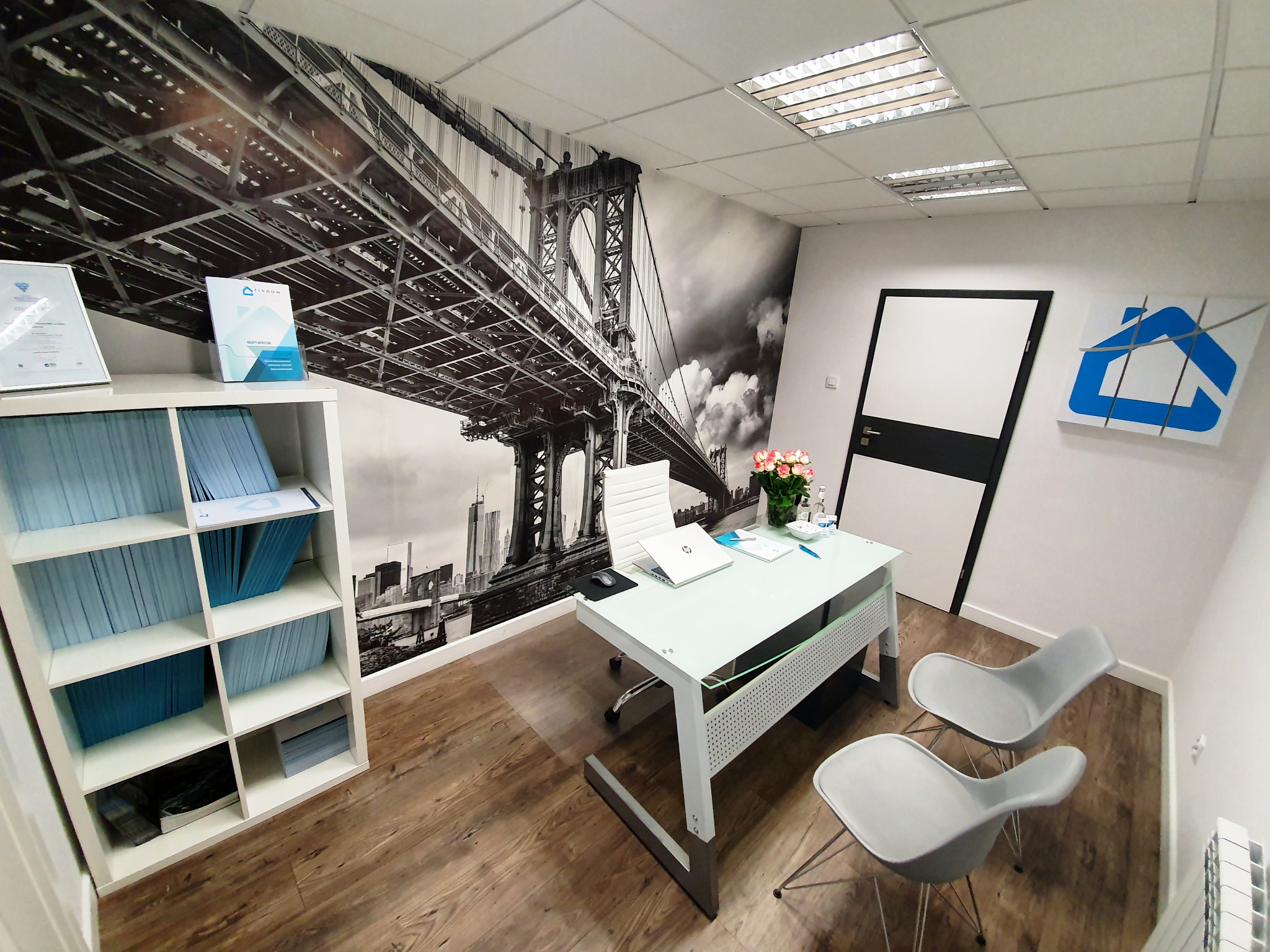 Nowoczesne biuro doradcy finansowego z widokiem na most na fototapecie, białe meble, dokumenty na szklanym biurku i logo firmy z niebieskim domem.