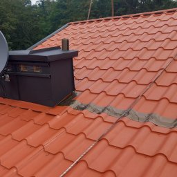 TomAlpin - Czerwona dachówka ceramiczna z czarnym kominem wentylacyjnym i anteną satelitarną. Widoczne uszczelnienie taśmą dekarską. Tło stanowią zielone drzewa.