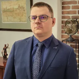 Portret mężczyzny w okularach i granatowym garniturze na tle biura z ceglaną ścianą, globusem i figurką Temidy.