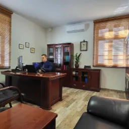 Wnętrze gabinetu: mężczyzna w okularach i niebieskiej marynarce siedzi za dużym, mahoniowym biurkiem, pracując na laptopie; w tle regał z książkami, zegar ścienny i żaluzje w oknach.