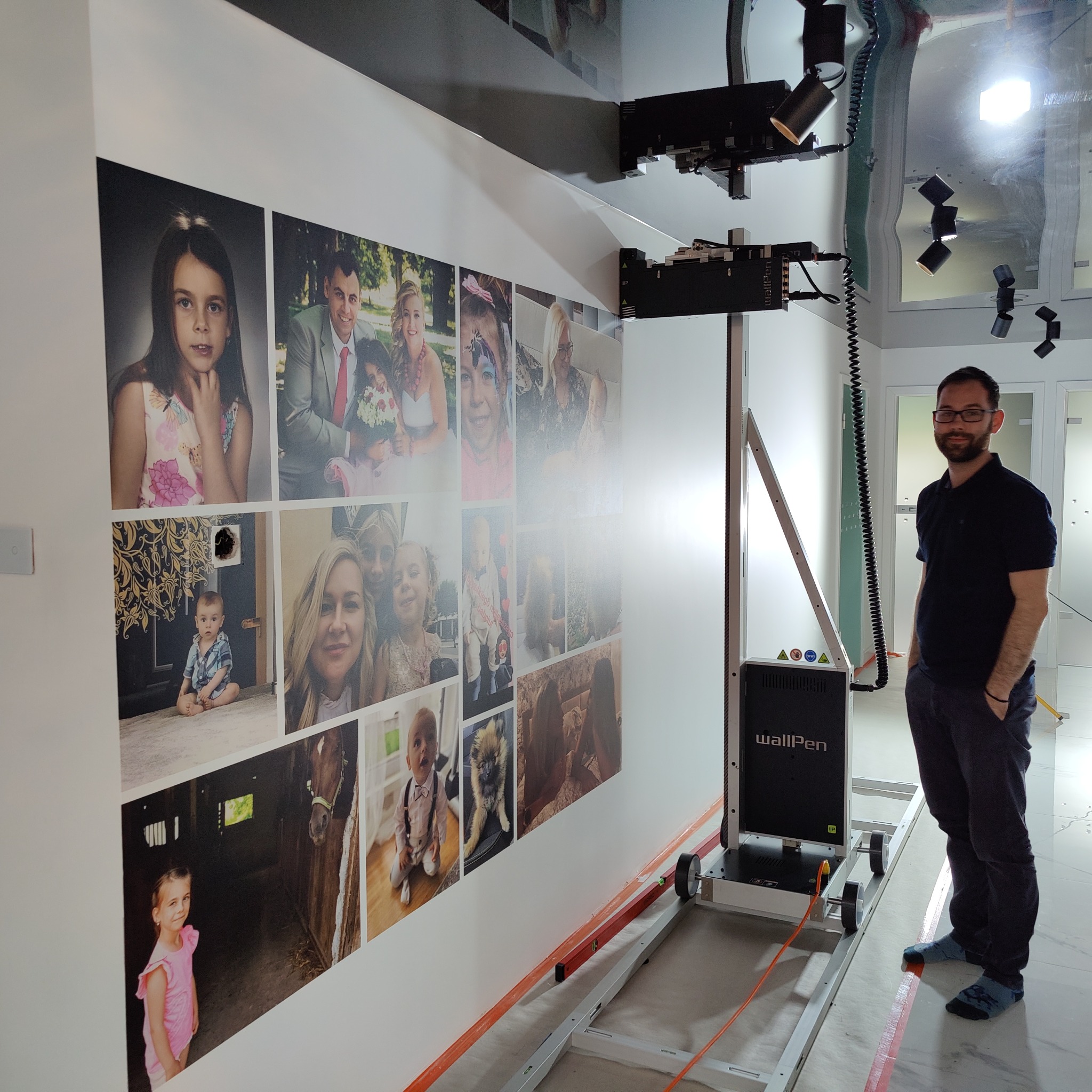 Ściana ozdobiona kolażem fotografii portretowych drukowanych za pomocą urządzenia WallPen, obok stoi mężczyzna z rękami w kieszeniach.