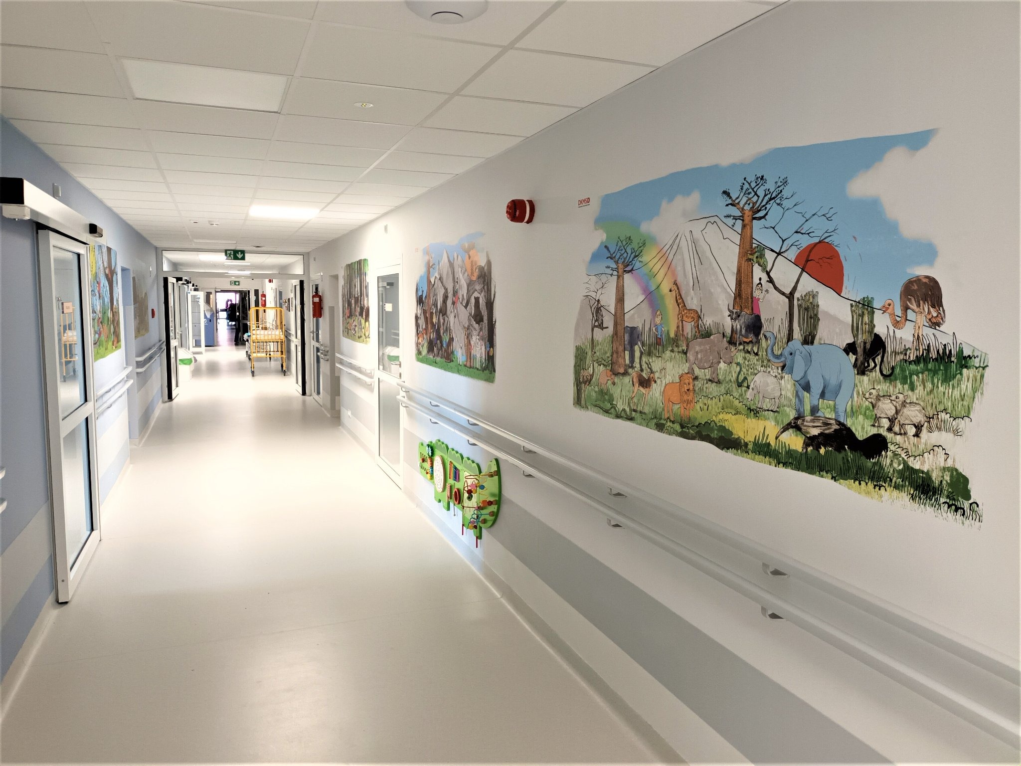Korytarz szpitalny z malowidłami ściennymi przedstawiającymi afrykańskie zwierzęta i krajobrazy, dodatkowo zabawki edukacyjne na ścianie.