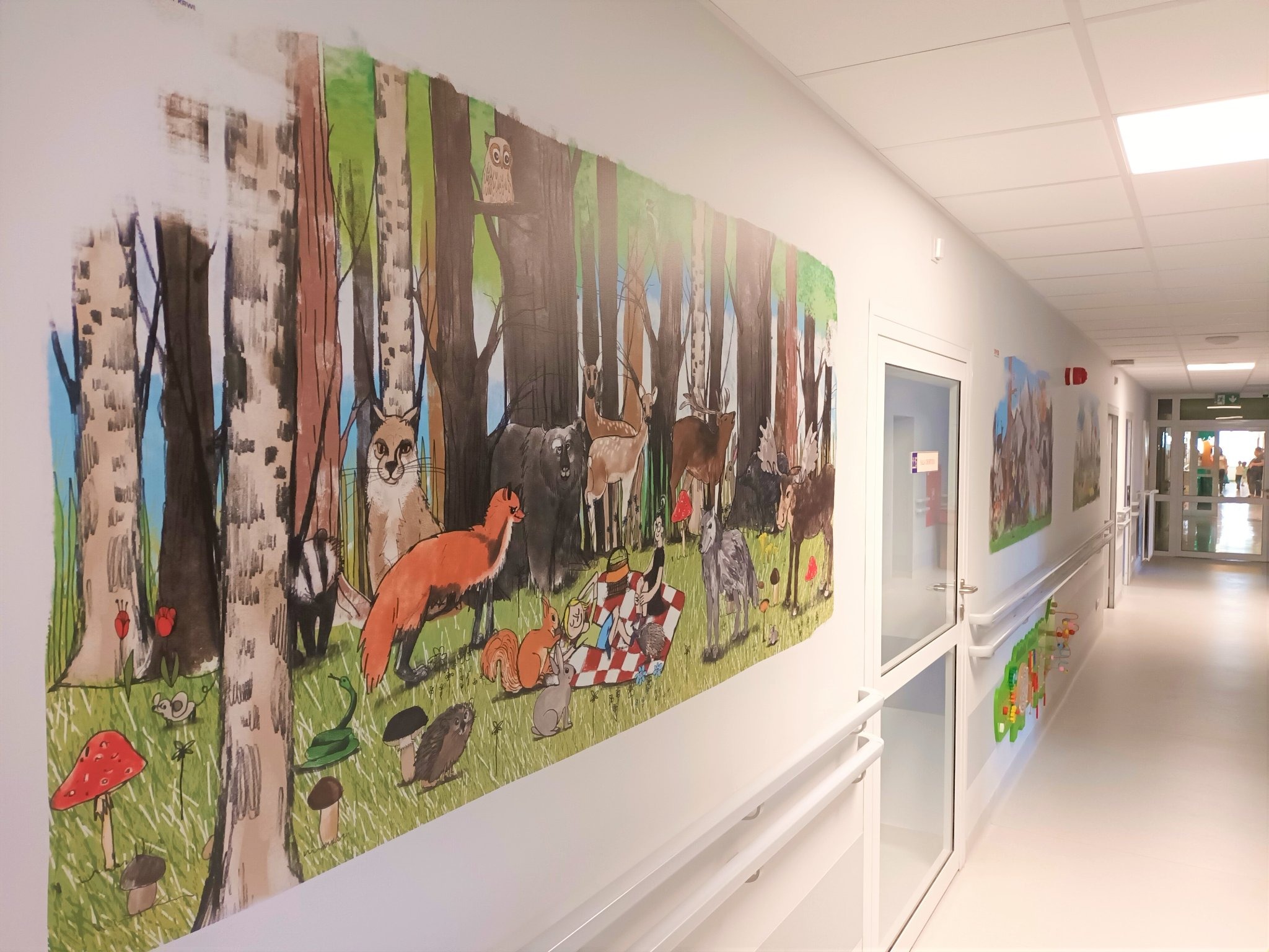 Ilustracja na ścianie korytarza przedstawiająca leśny krajobraz z różnymi zwierzętami, takimi jak lis, jeleń, niedźwiedź i sowa, stylizowana na rysunek dla dzieci, z elementami pikniku na trawie.