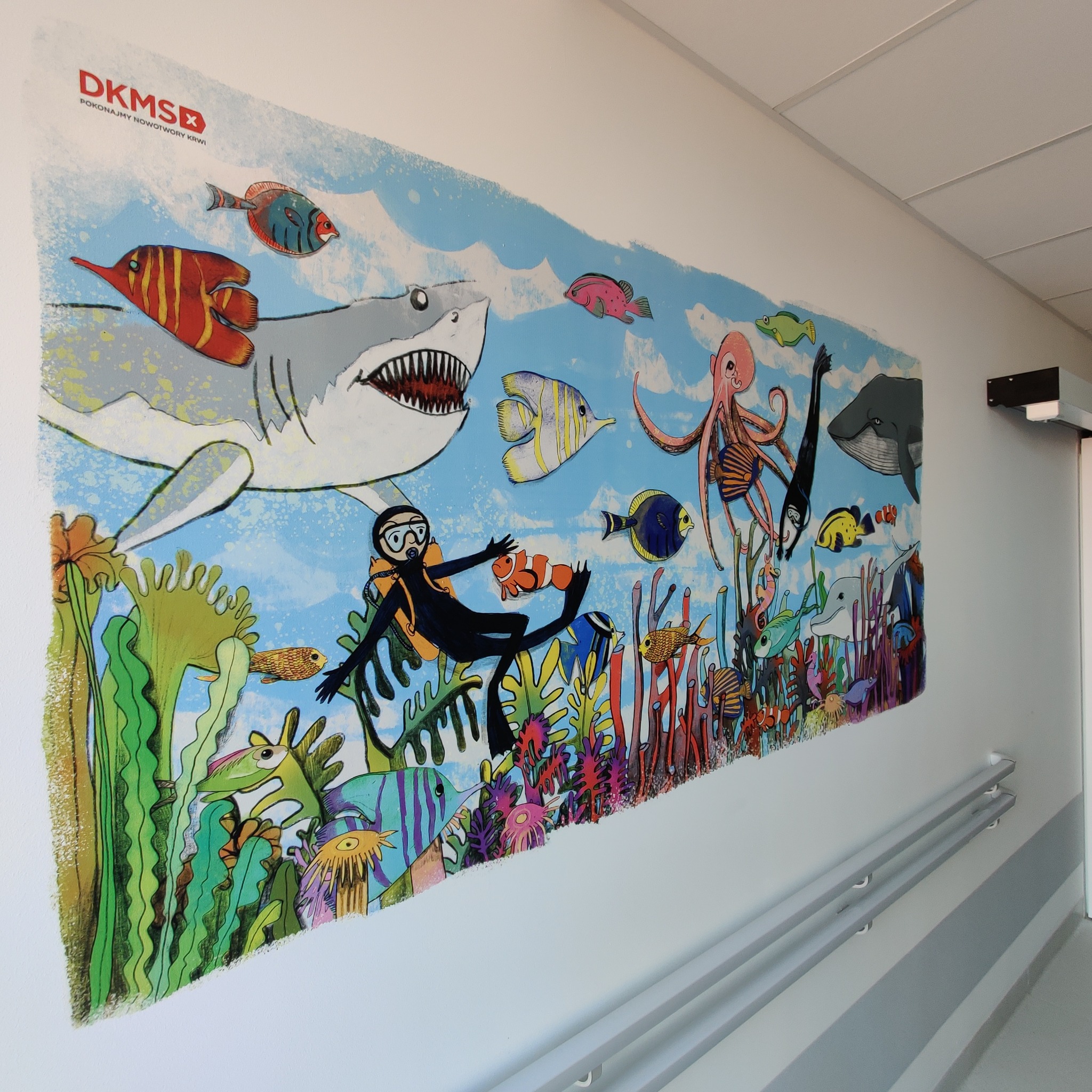 Kolorowa ilustracja podwodnego świata z rekinem, nurkiem, ośmiornicą i rybami na ścianie korytarza szpitalnego, z logo DKMS w lewym górnym rogu.