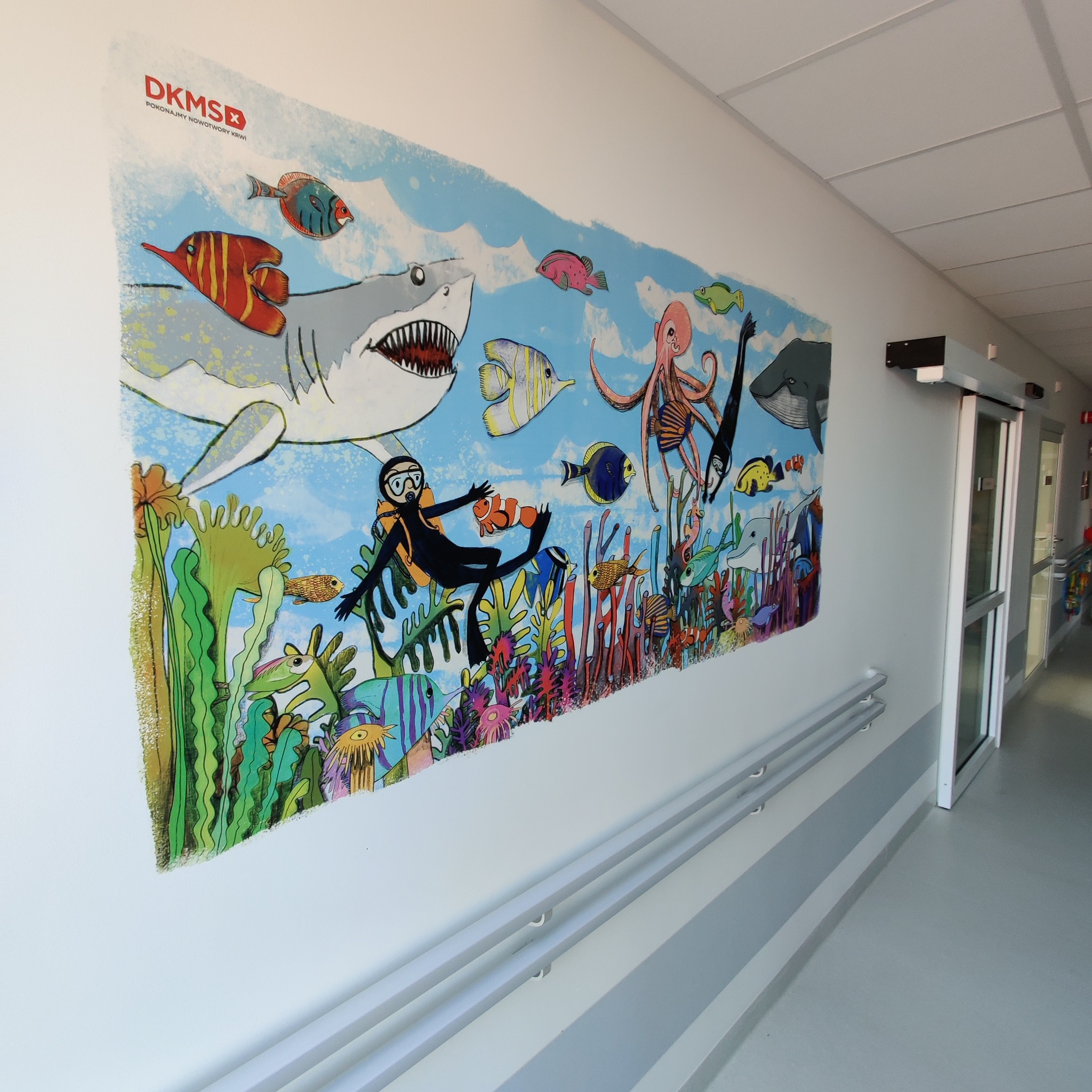 Ilustracja na ścianie w szpitalnym korytarzu przedstawiająca podwodny świat z rekinami, nurkiem i różnymi rybami, wykonana w stylu dziecięcym z logo DKMS.