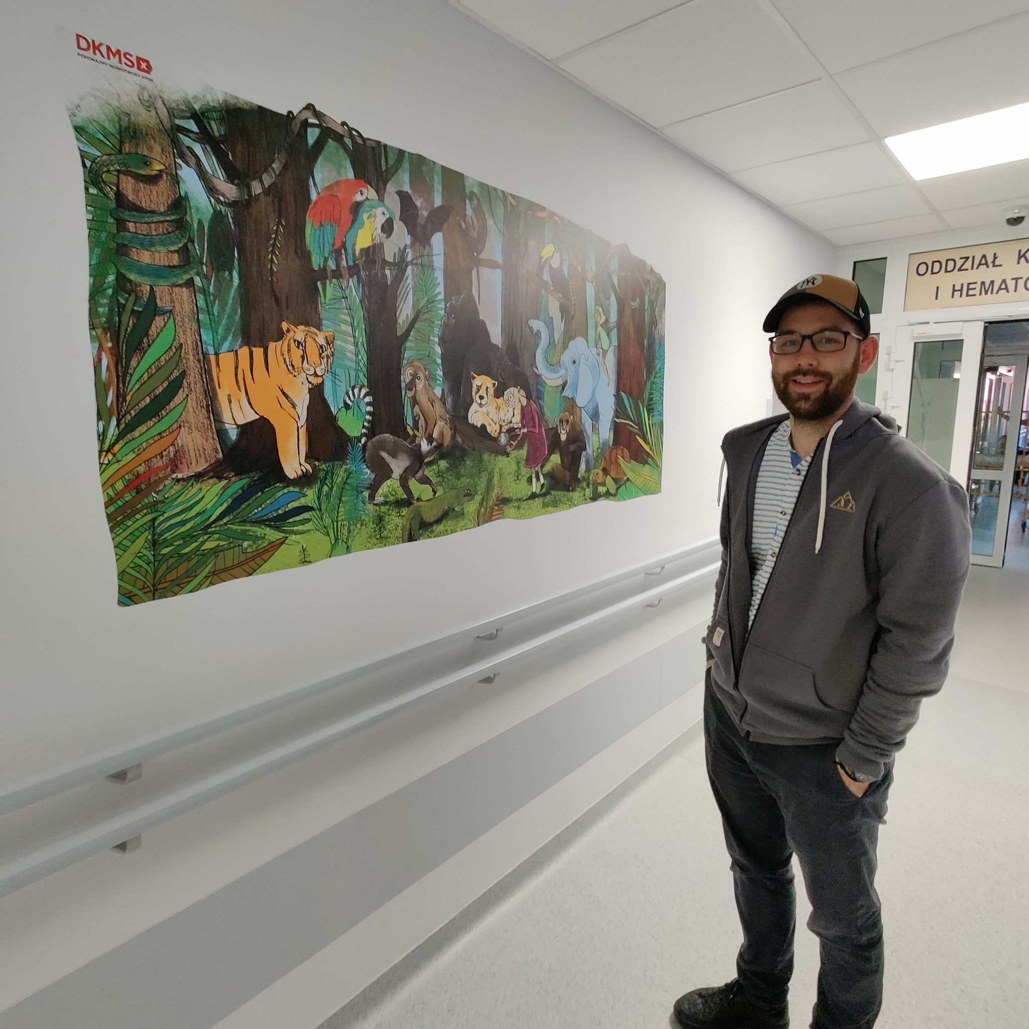 Mężczyzna pozuje na tle ściany szpitalnego korytarza z kolorową ilustracją dżungli z tygrysem, małpami, słoniem i papugami, wykonaną na zlecenie fundacji DKMS.