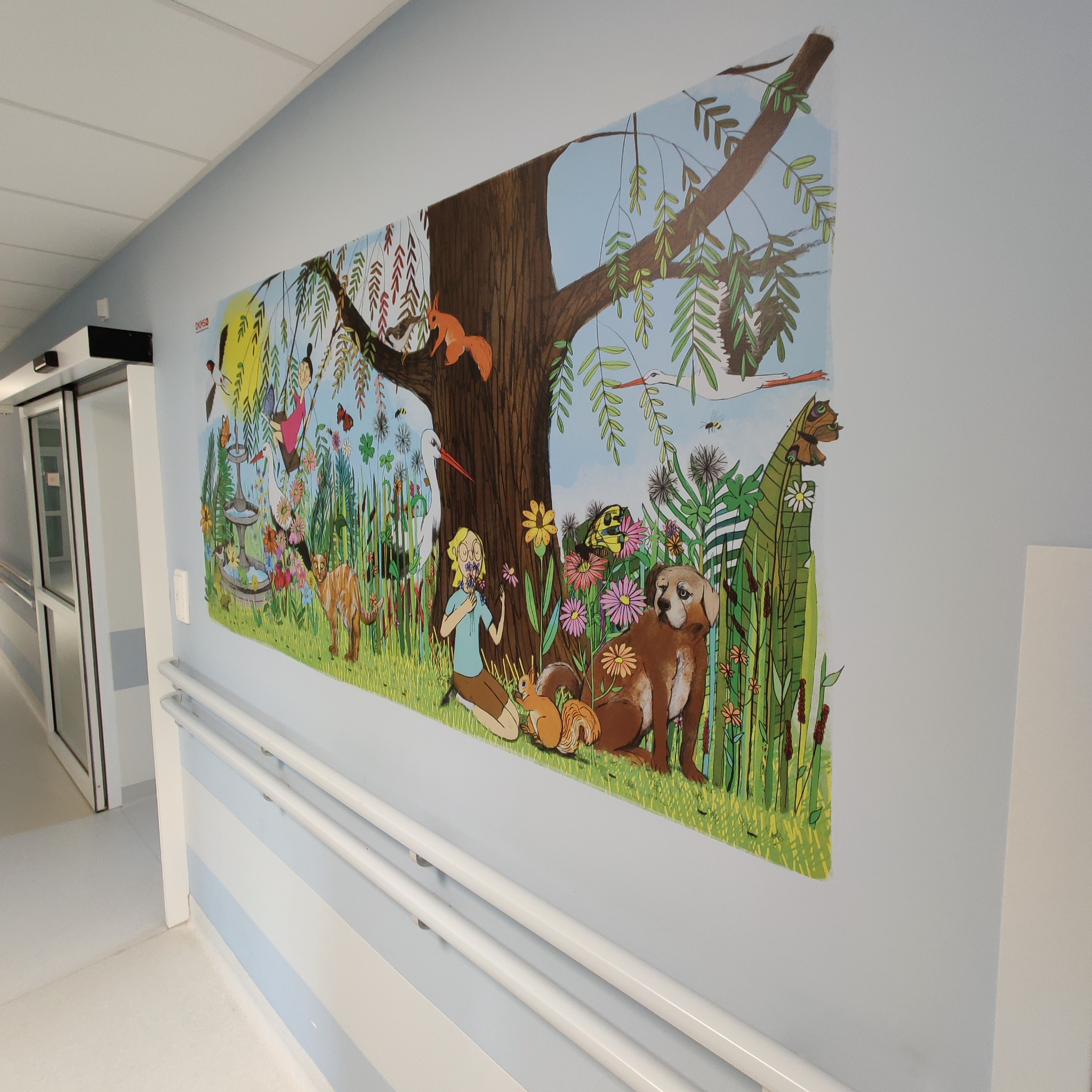 Ilustracja na ścianie w szpitalnym korytarzu przedstawiająca dziewczynkę, zwierzęta i rośliny w stylu dziecięcym.
