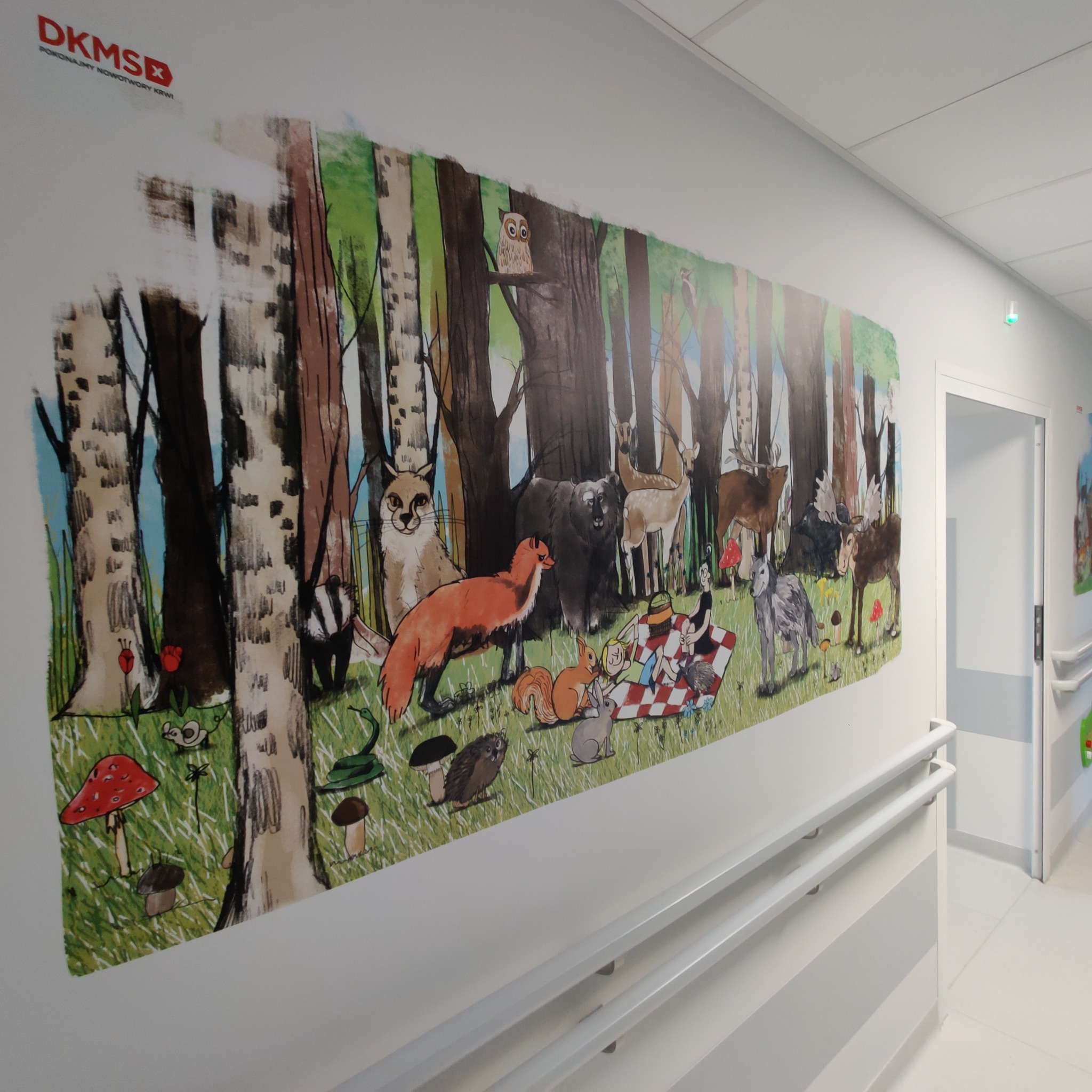 Ilustracja na ścianie w szpitalnym korytarzu przedstawiająca leśny krajobraz z zwierzętami i piknikiem, stylizowana na rysunek dla dzieci.