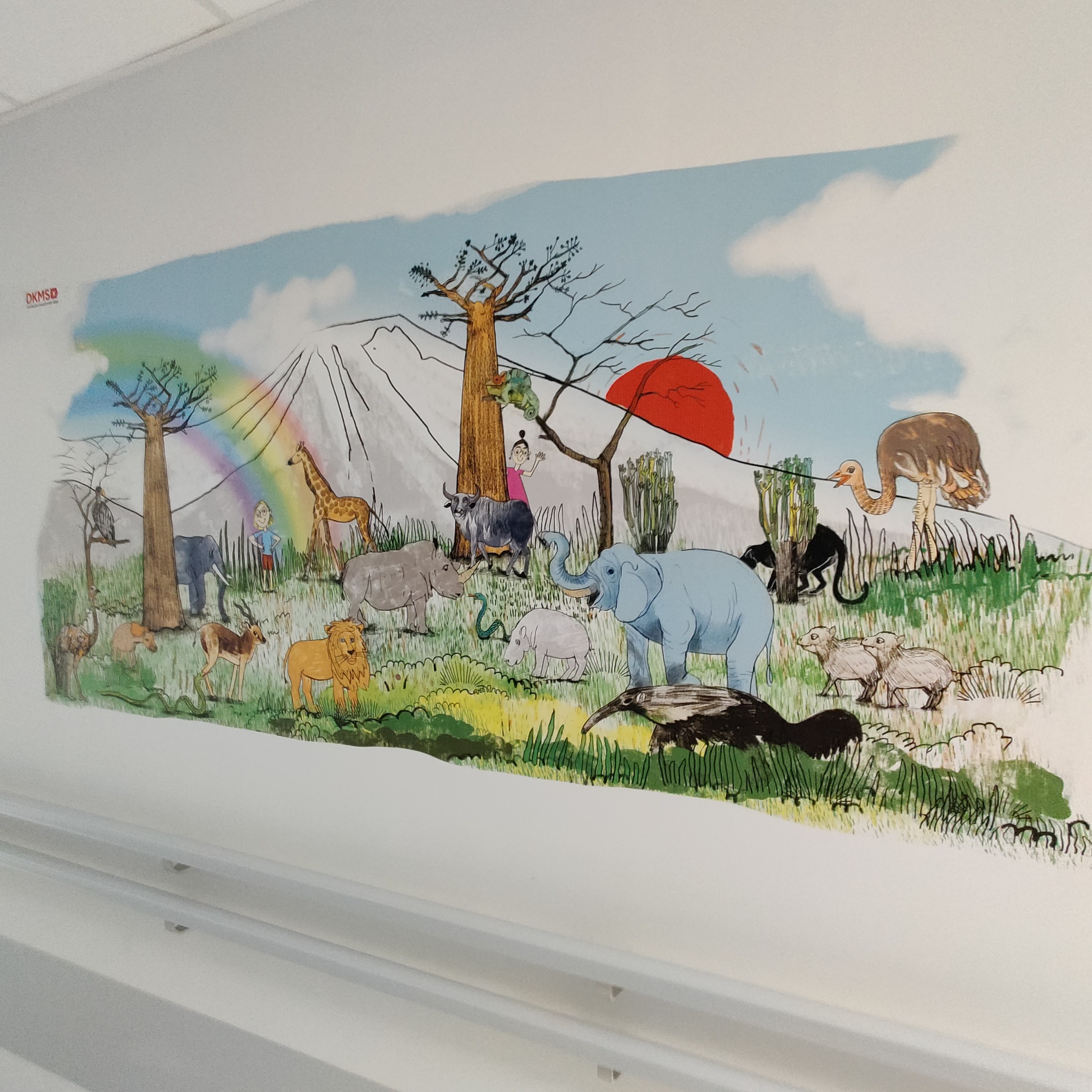 Ręcznie malowana scena afrykańskiej sawanny z różnorodnymi zwierzętami, w tym słoniem, żyrafą, lwem i strusiem, umieszczona na ścianie, widoczna z perspektywy dolnego kąta.