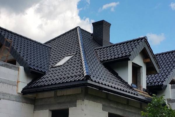Nowy, czarny dach z ceramicznej dachówki, widoczny z dołu, z oknem dachowym i kominem, na tle błękitnego nieba z chmurami, w trakcie budowy domu z białego pustaka.