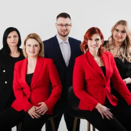 Portret grupowy pięciu osób w eleganckich strojach: trzech kobiet w czarnych marynarkach i dwóch w czerwonych, oraz mężczyzny w garniturze w kratę, na jasnym tle.