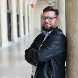 Portret adwokata w okularach, oparty o kamienną kolumnę z rękami założonymi na piersiach, w tle arkady i oświetlenie uliczne.