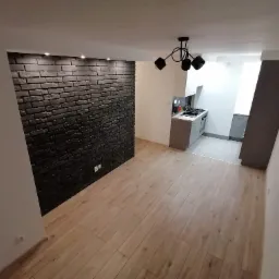 Salon z dekoracyjną ścianą z czarnej cegły, oświetleniem punktowym i podłogą z paneli drewnopodobnych, widok na aneks kuchenny z szarymi szafkami.