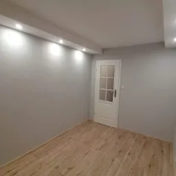 Pomalowane na szaro wnętrze pokoju z ułożonymi panelami podłogowymi w odcieniach drewna i białą, przeszkloną częściowo ścianką drzwiową.