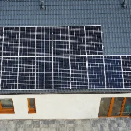 Widok z góry na dom z ciemną dachówką i panelami słonecznymi, częściowo zacieniony dach, kostka brukowa wokół domu.