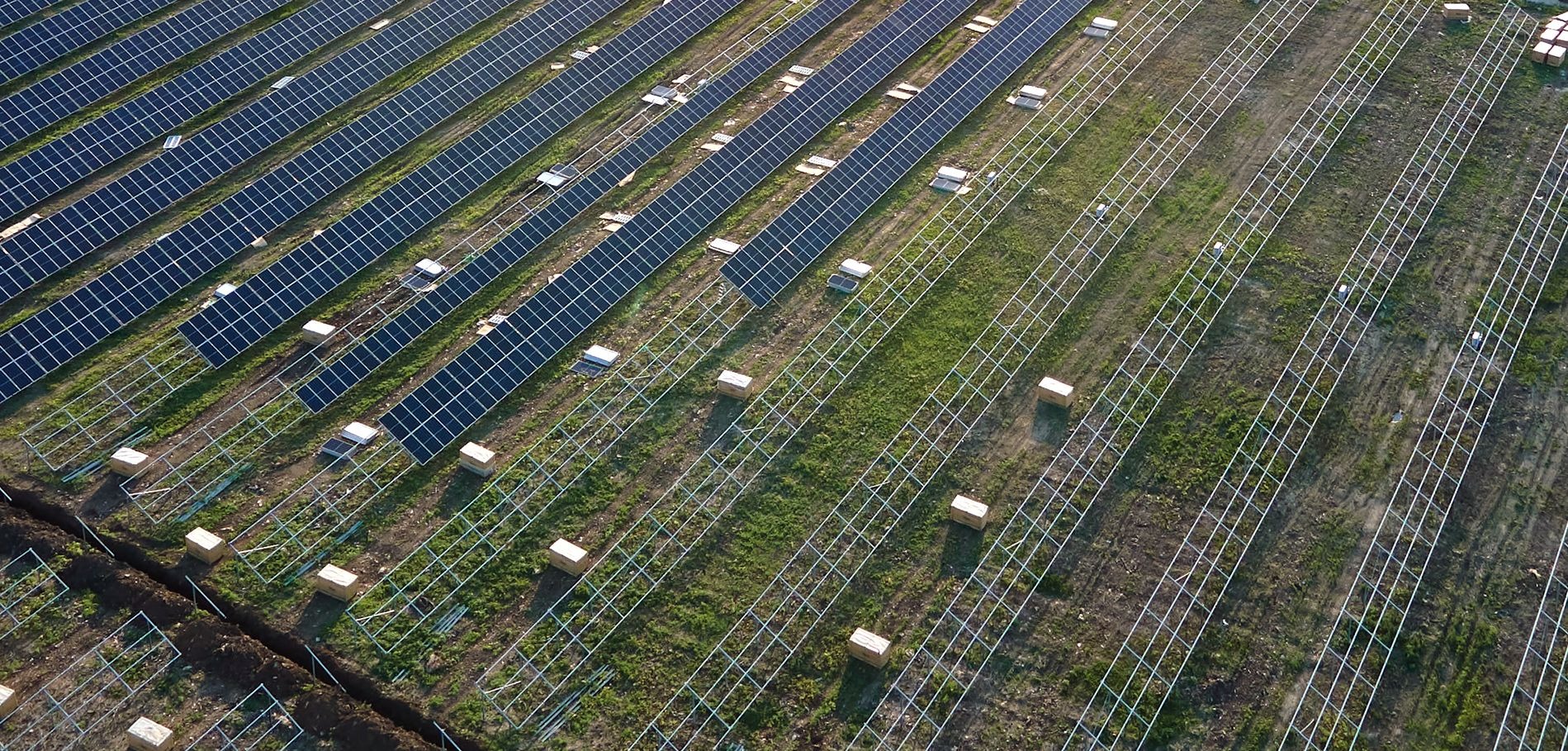 Widok z drona na pole z częściowo zainstalowanymi panelami słonecznymi i konstrukcją wsporczą, widoczny wykop pod instalację elektryczną.